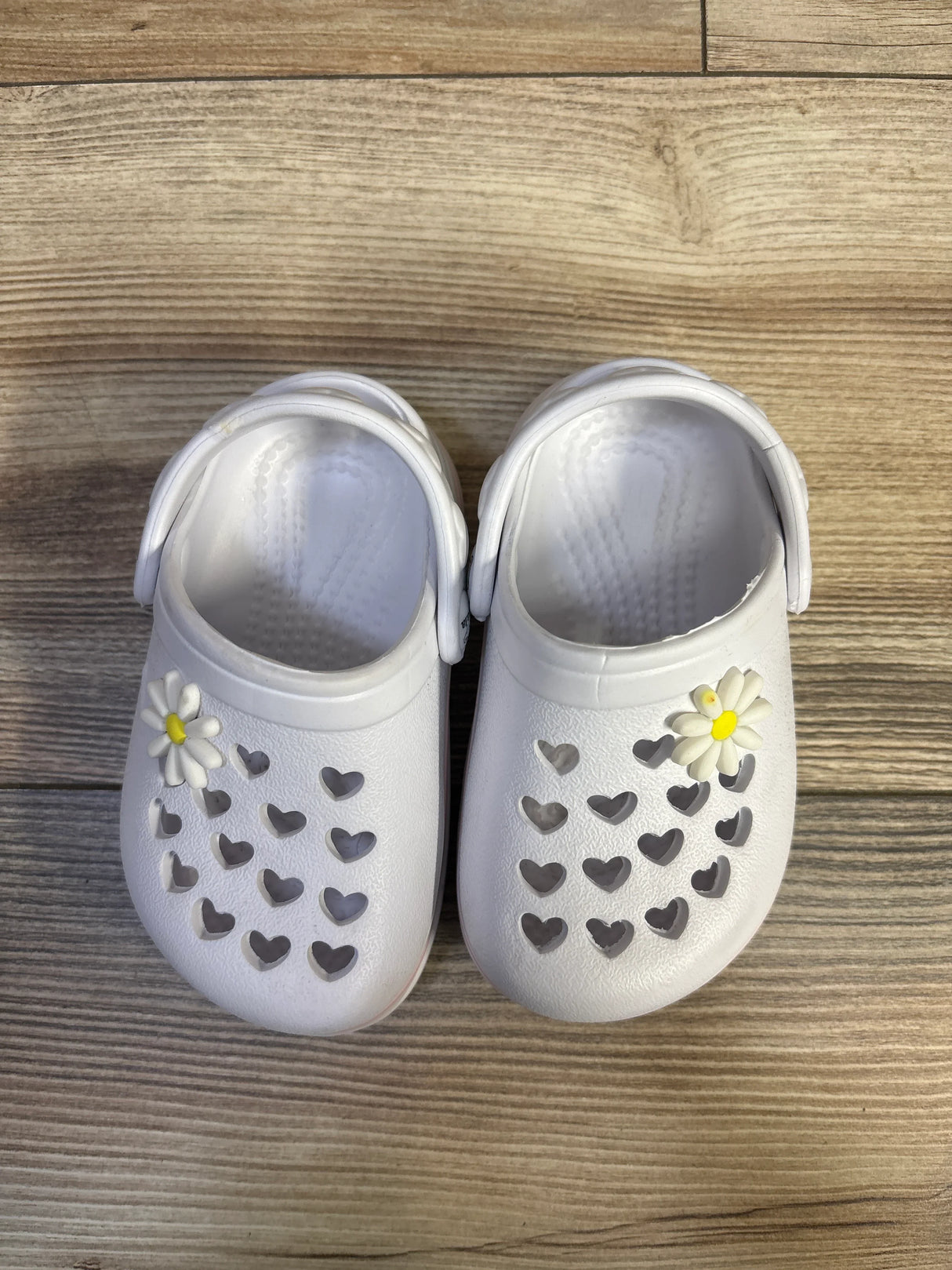Overhead view of white Koala Kids clogs showing heart-shaped cutouts and daisy charms on a wood floor
