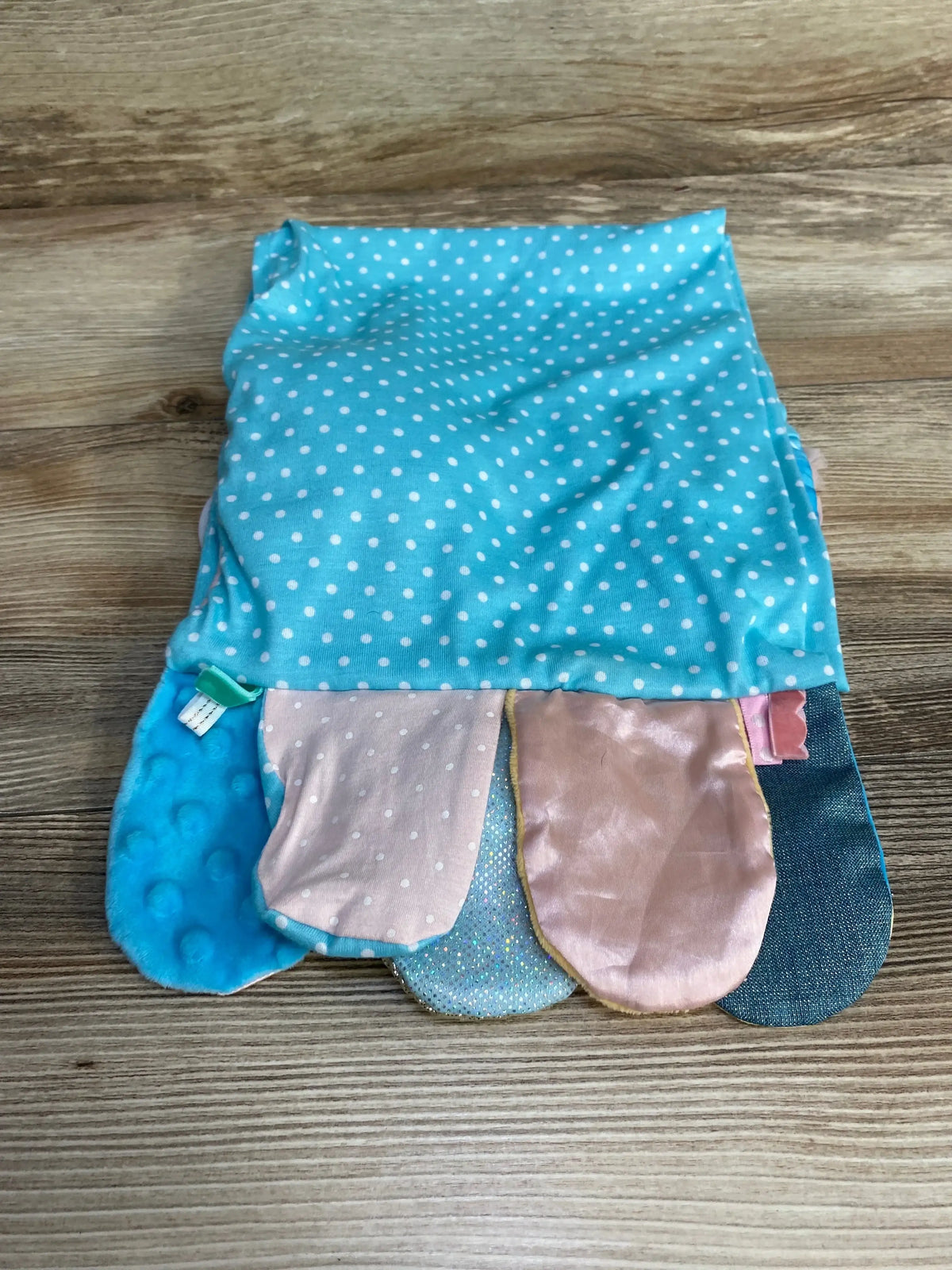 Top-down of Demdaco Activity Scarf (Mermaid) showing blue polka-dot fabric and five textured sensory tabs on wooden surface