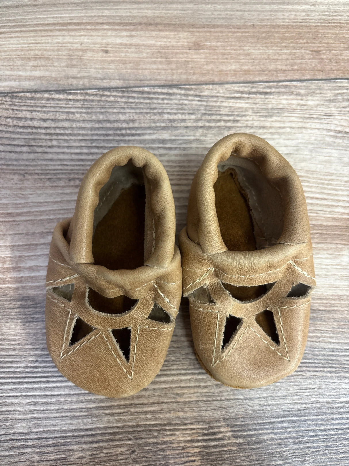 Top view of newborn tan leather moccasins with star cutout toes, stitched edges and elasticized cuffs on gray wood background