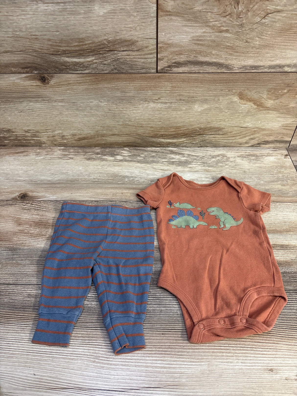 Flat lay of 2pc baby set: rust-orange bodysuit with green dinosaur print and blue-gray striped pants on wood background