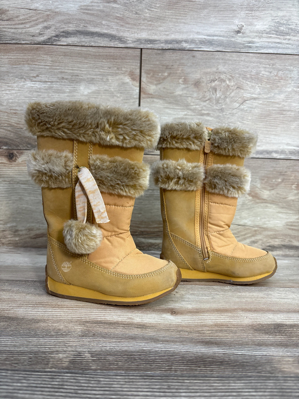 Tan fur-lined Timberland kids winter boots with pom-poms on wooden floor background