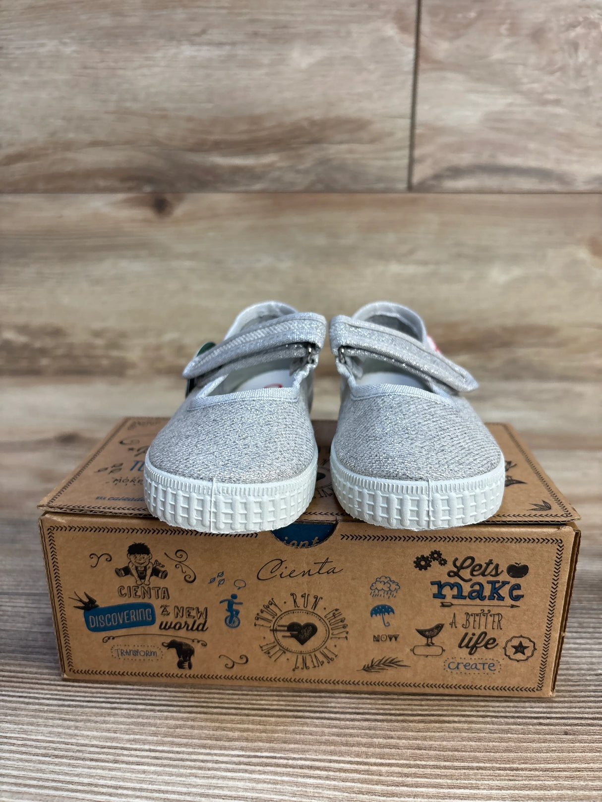 Front view of toddler Mary Jane flats size 7C, silver glitter upper with Velcro strap and white rubber sole on Cienta shoebox, wood background