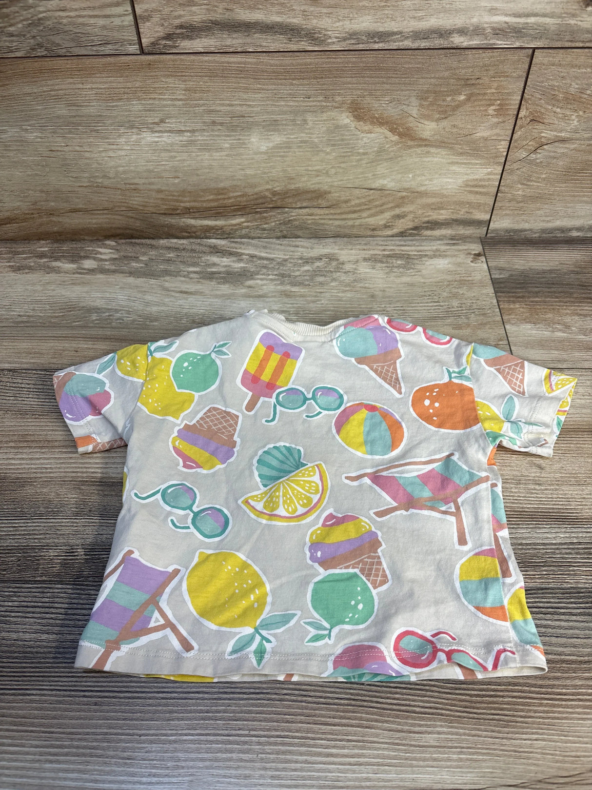 Flat-lay of H&M short-sleeve baby shirt (cream, 9m) showing colorful summer prints, including lemons, ice cream cones, and sunglasses, on wood background