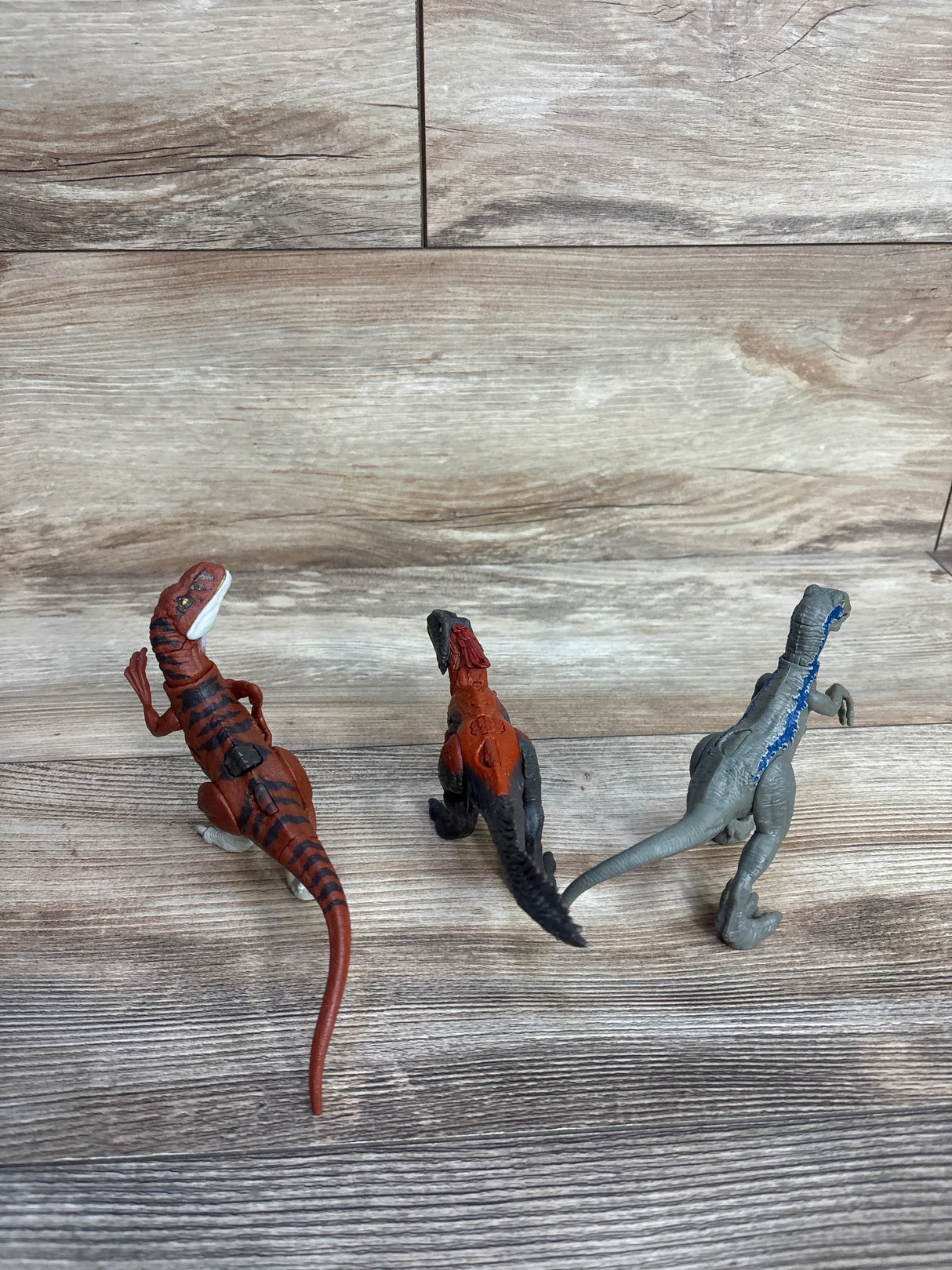 Three plastic dinosaur figurines on a wooden floor background, viewed from behind.
