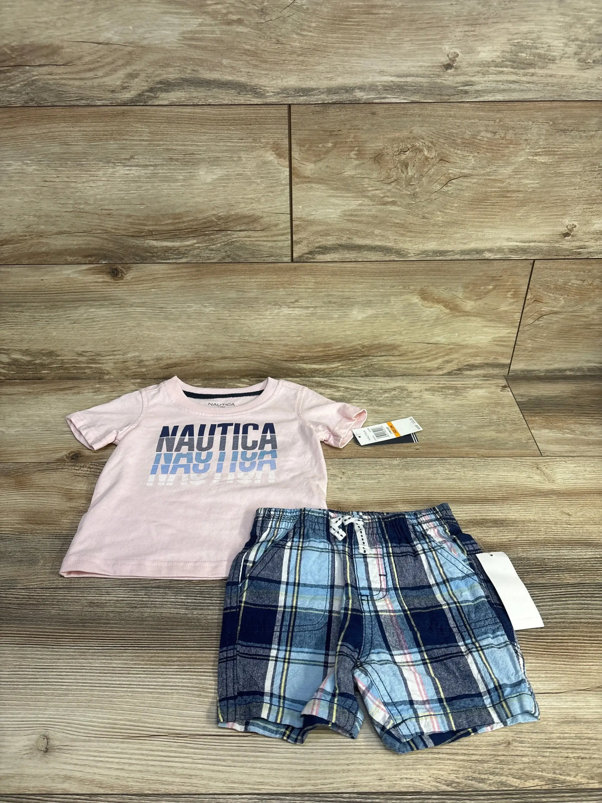 Flat lay of Nautica baby set: pink T-shirt with NAUTICA logo and blue plaid drawstring shorts on wood background