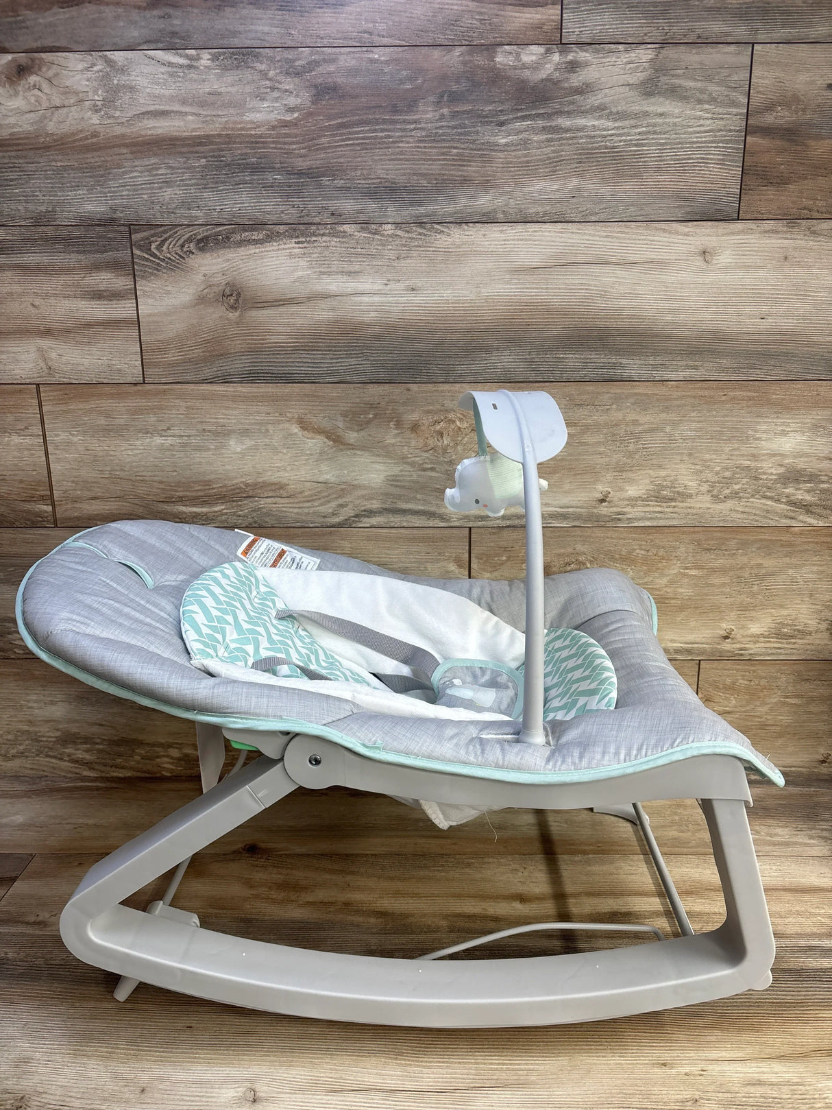 Side view of Ingenuity Keep Cozy 3-in-1 bouncer with grey fabric, mint insert, rocker base and toy bar on wood background