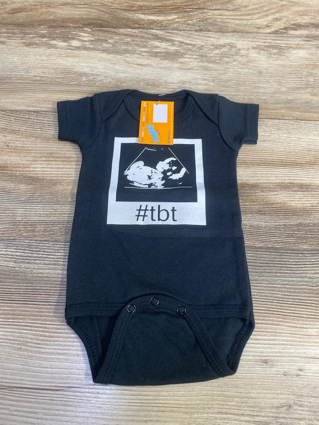 Flat lay of black infant bodysuit (size 0-6 months) showcasing white sonogram graphic and 'tbt' text on a light wood background