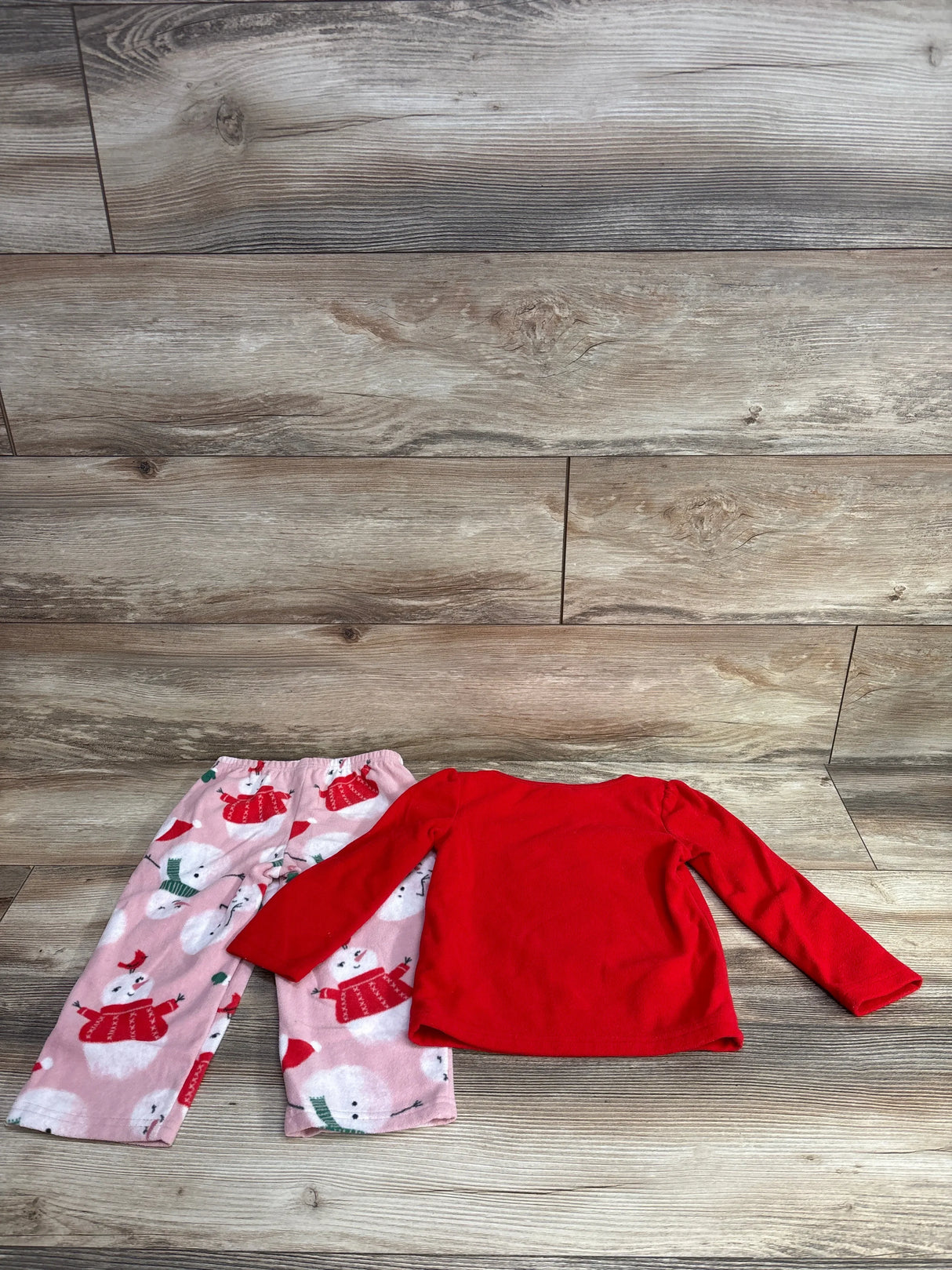 Children's red long-sleeve shirt and pink snowman print pants laid on wooden floor, kids holiday pajama set