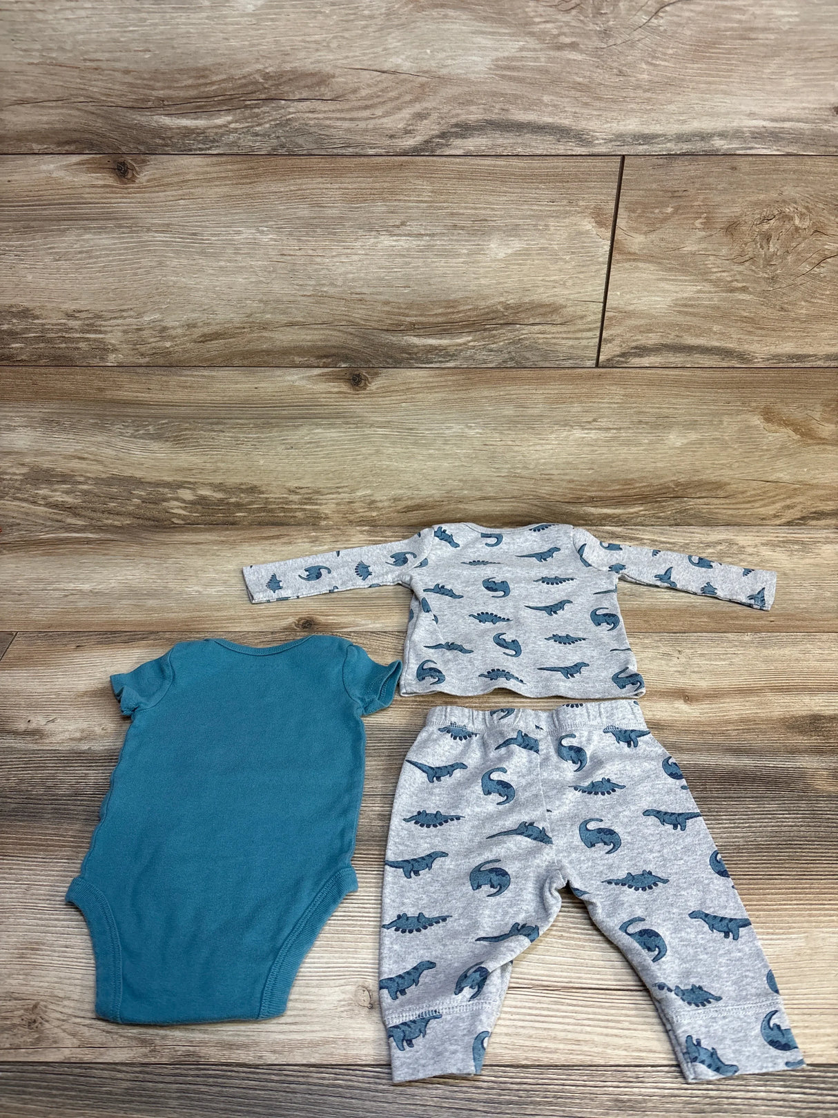 Flat lay of 3-piece dinosaur set size 3M showing teal bodysuit and gray dinosaur-print long-sleeve top and matching pants on wood floor
