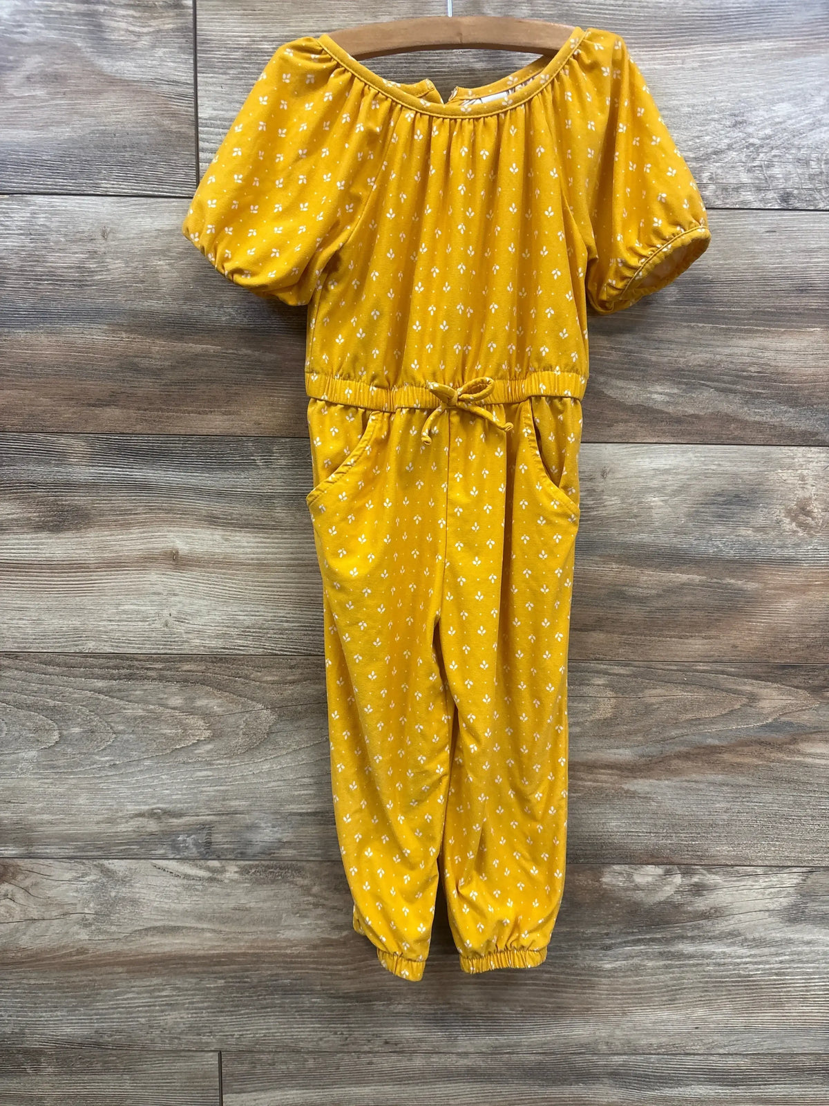 Front view of Old Navy yellow jumpsuit with puff sleeves, drawstring waist, pockets and elastic ankles on wood background