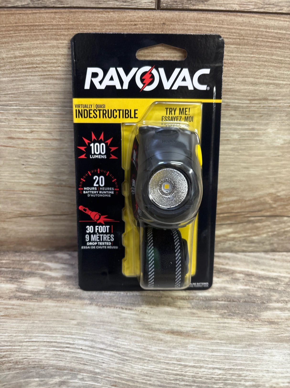 Front view of Rayovac Virtually Indestructible LED headlamp in retail blister pack showing lens and elastic headband, packaging text 100 lumens and 20 hours, wooden background