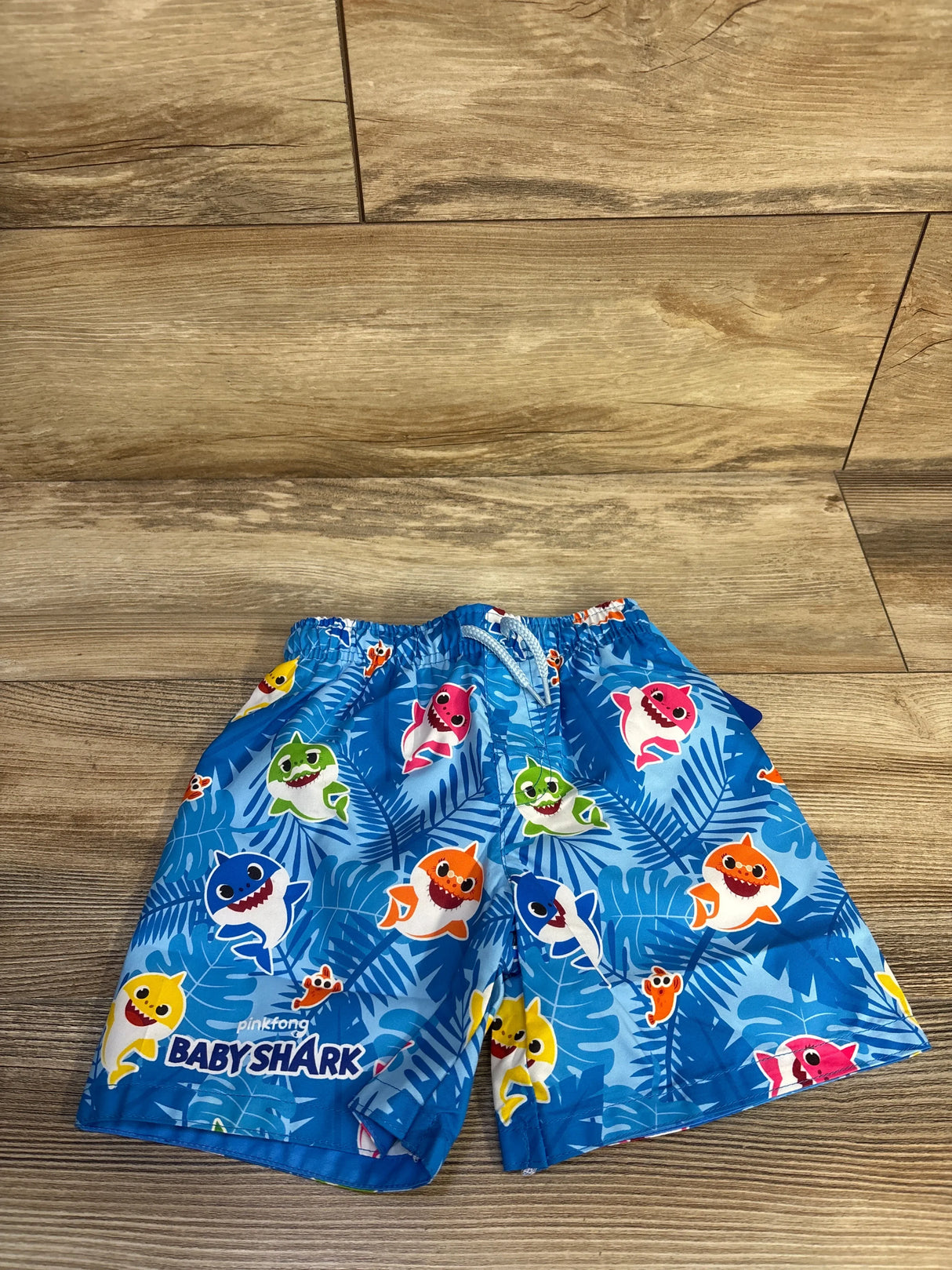 Flat lay of blue Baby Shark swim trunks showing elastic waistband and colorful shark characters on tropical-leaf print on wood floor