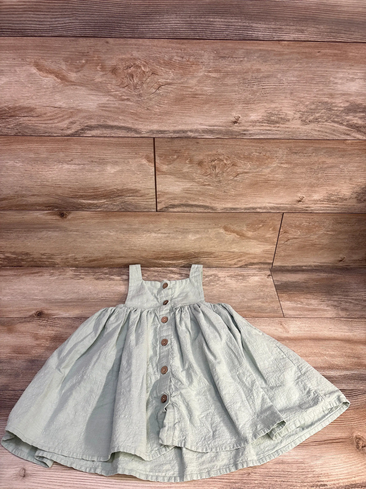 Front view of a green sleeveless button-front dress in size 2T showcasing its gathered skirt and brown buttons on a wood floor