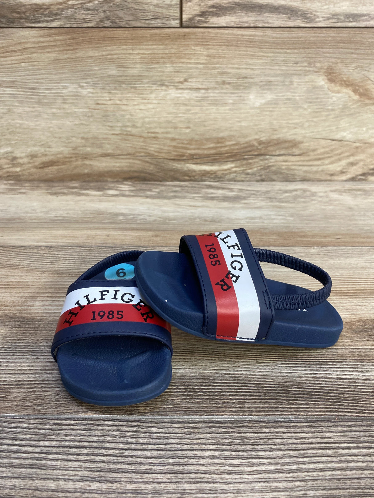 Navy blue toddler sandals with red and white Hilfiger 1985 straps on wood background