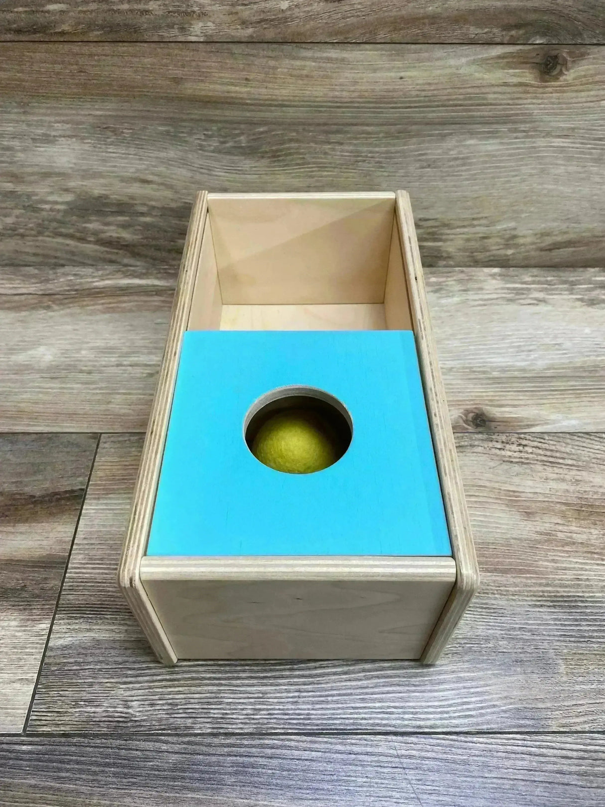 Top-down view of Lovevery Sliding Top Box showing turquoise sliding lid with round hole revealing yellow ball on wood floor background