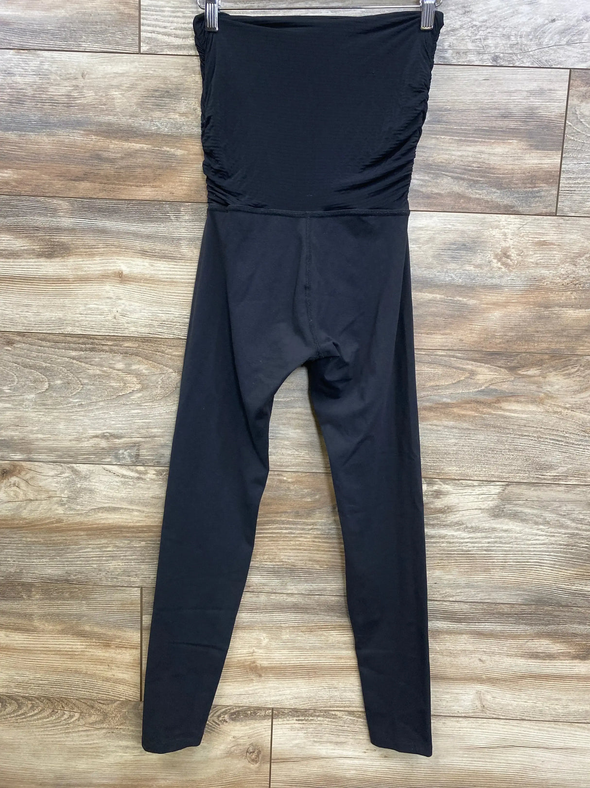 Front view of black maternity high-waist leggings showing over-the-bump tummy panel and ruched side detail on wood plank background