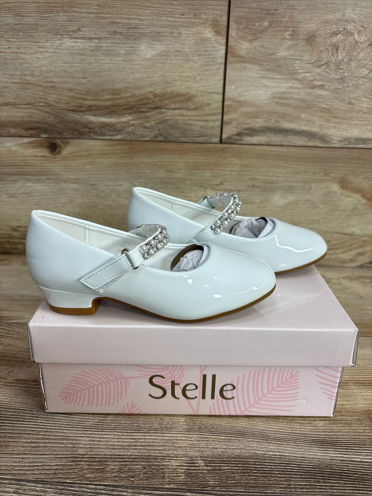 White patent Mary Jane shoes with rhinestone strap detail on a pink 'Stelle' shoebox, wooden background
