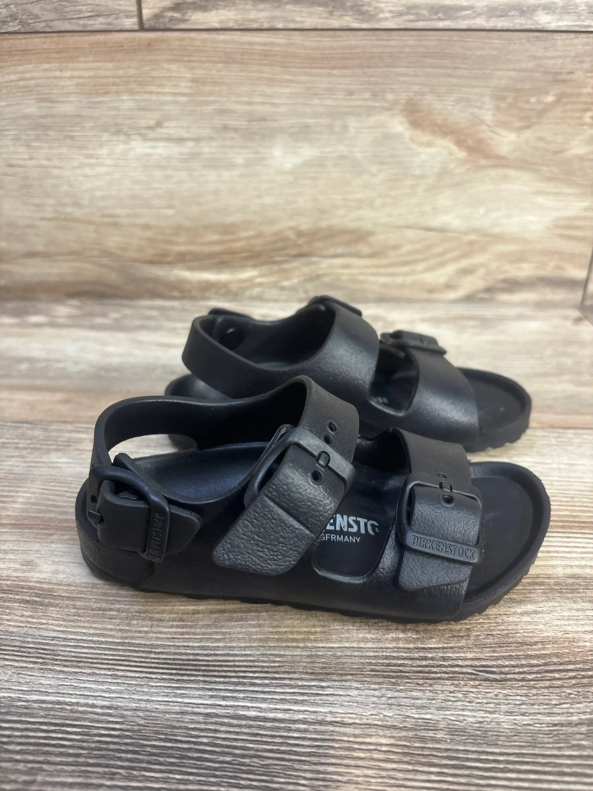 Front view of Birkenstock Milano EVA Sandals in black, showcasing three adjustable straps and backstraps on a wood-look floor
