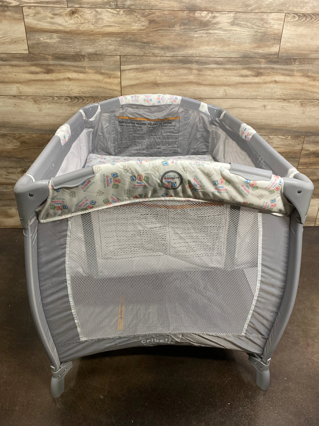 Front view of Cribs for Kids Cribette playard showing mesh sides and patterned padded top rail against wood wall