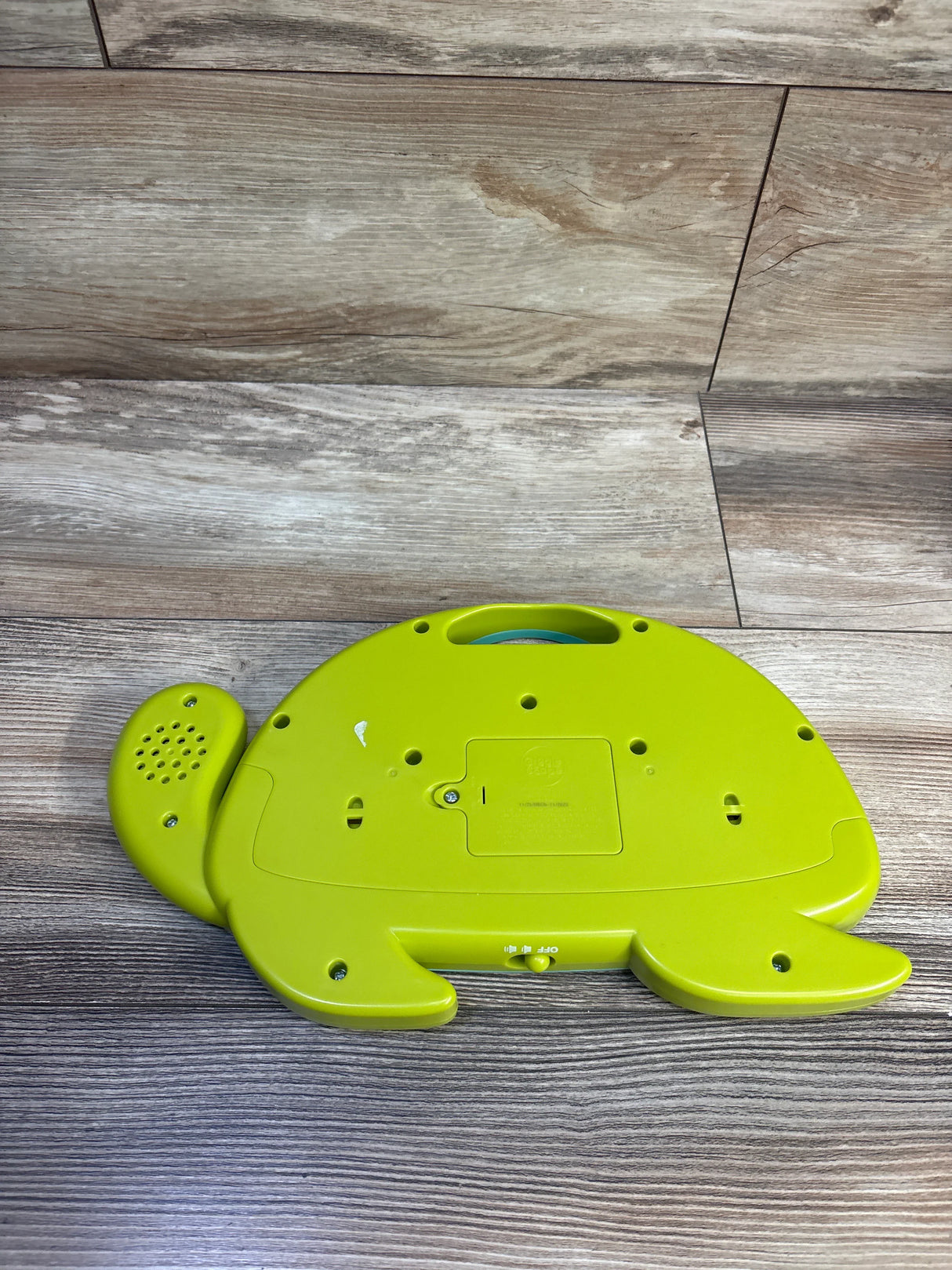 Underside of Gigglescape Turtle electronic drum showing battery compartment, speaker grille and on/off switch on wood floor