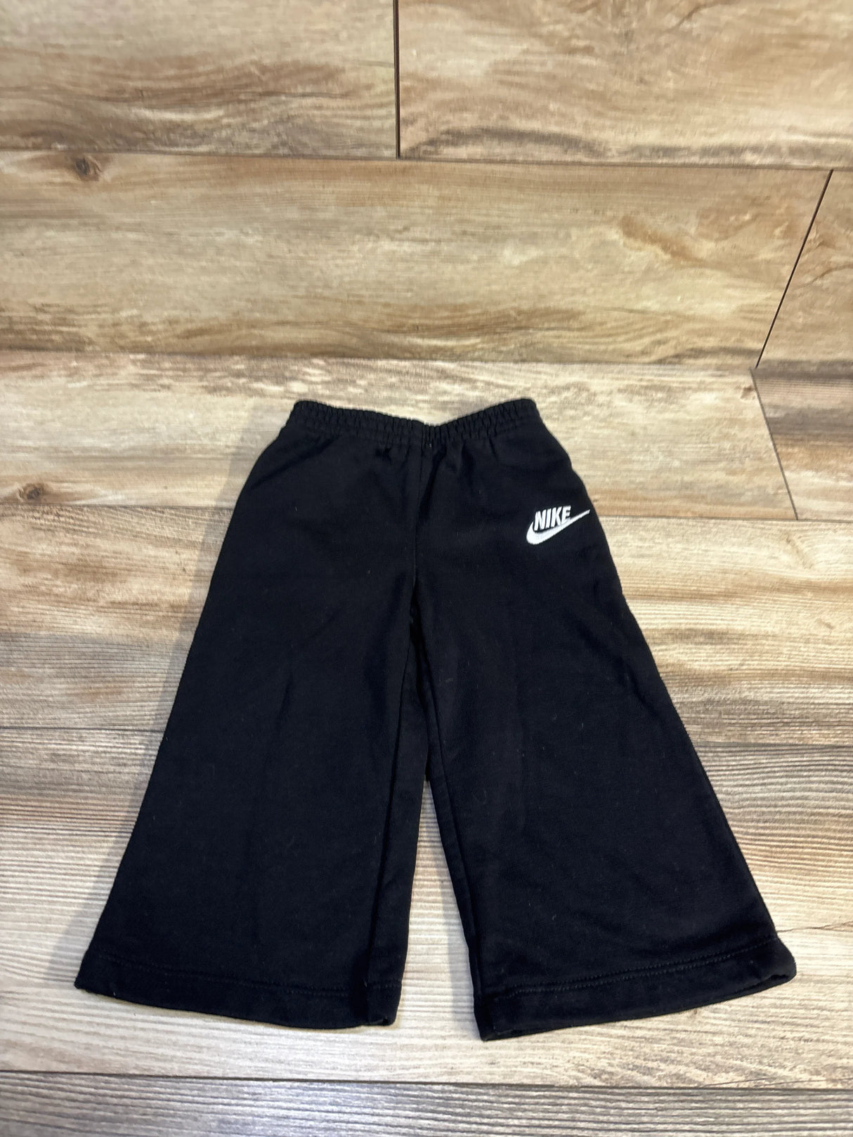 Front view of toddler black Nike wide-leg joggers showing elastic waistband and white swoosh logo on wood floor background