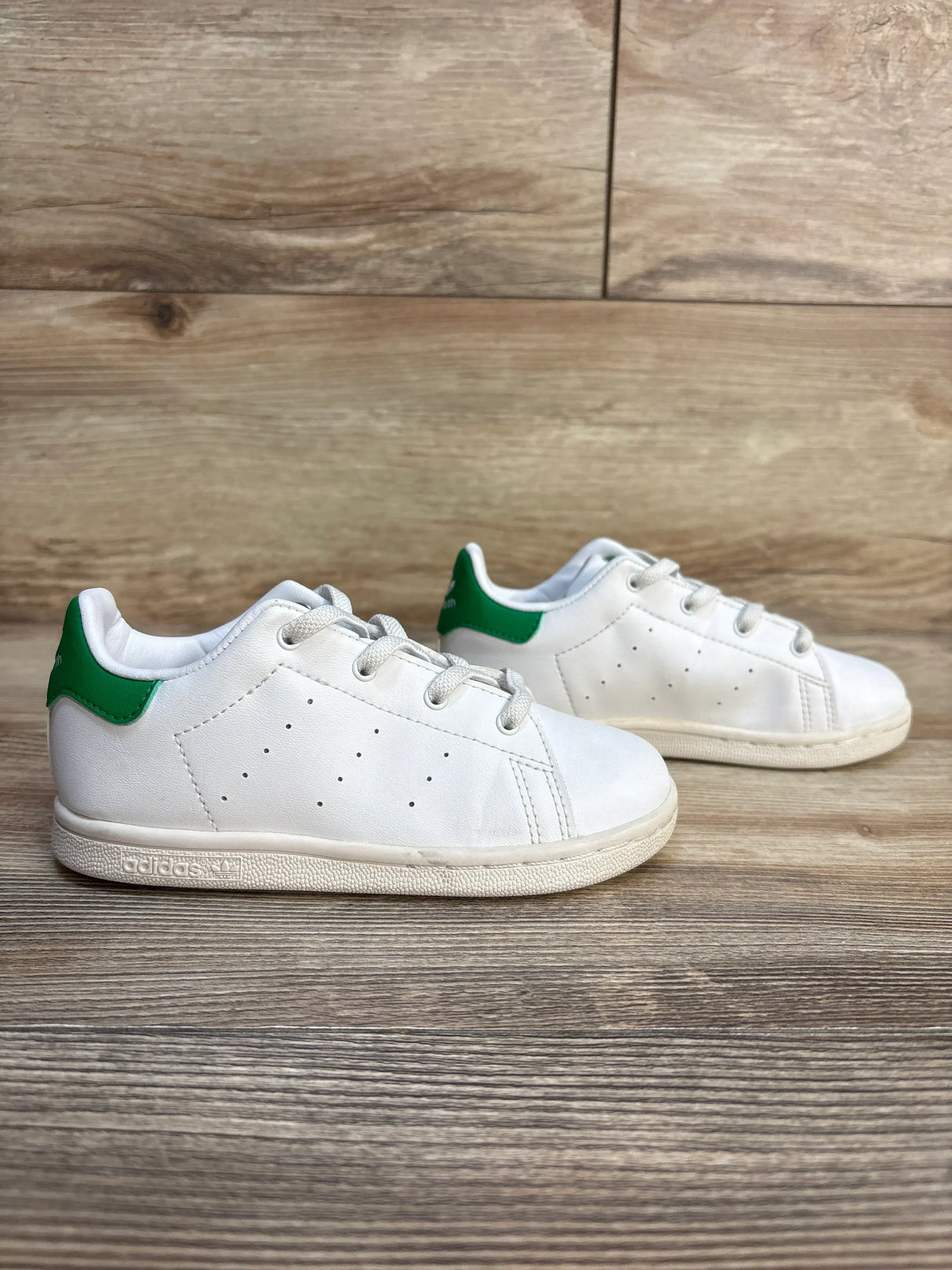 White kids' Adidas sneakers with green heel accents on wooden floor