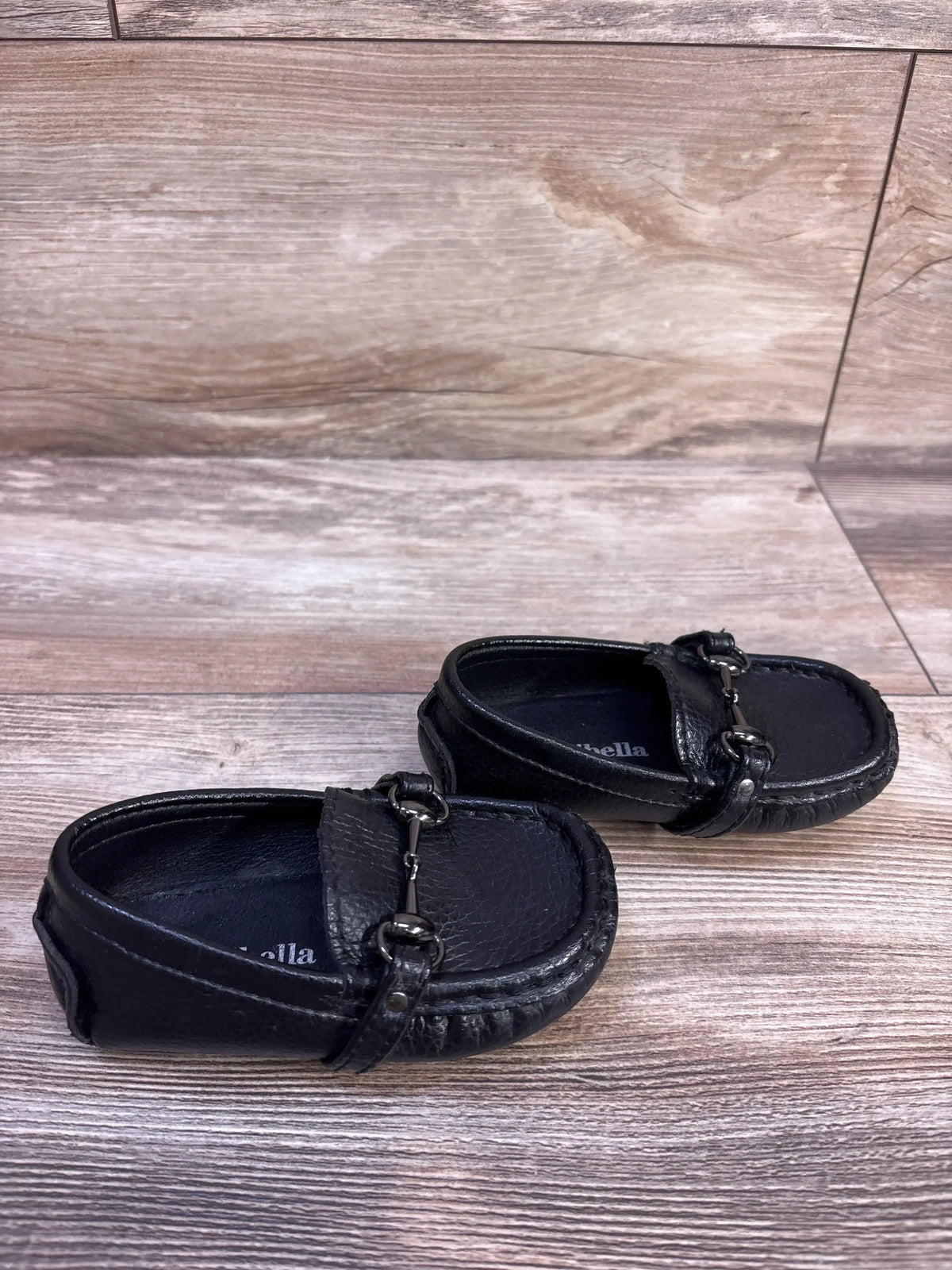 Angled top view of Minibella slip-on boat shoes with black leather, metal horsebit and stitching on light wood background