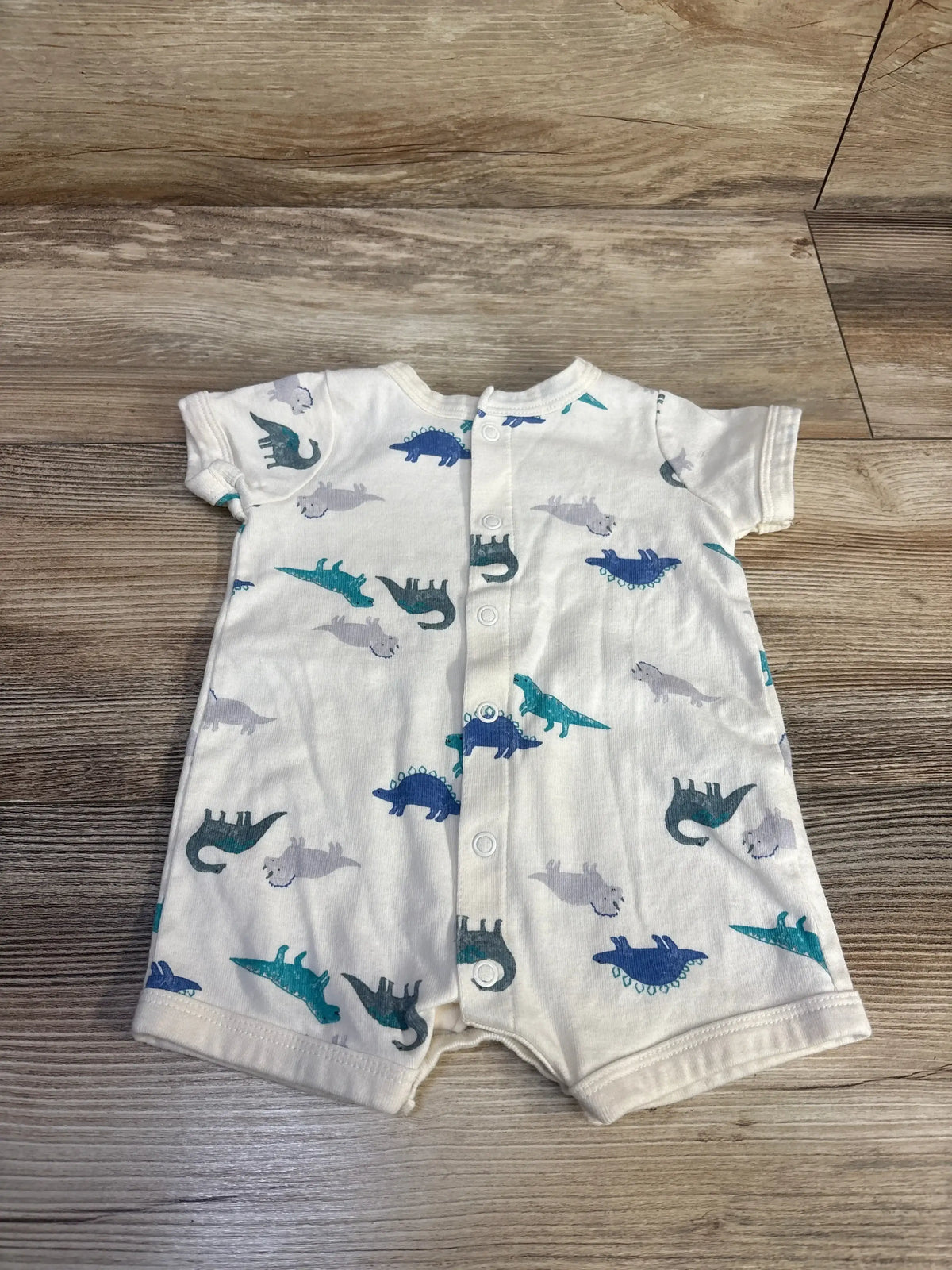 Flat lay of Carter’s Dinosaur Shortie Romper white sz 3m showcasing blue and grey dinosaur print short sleeves and front snap placket on wood plank background