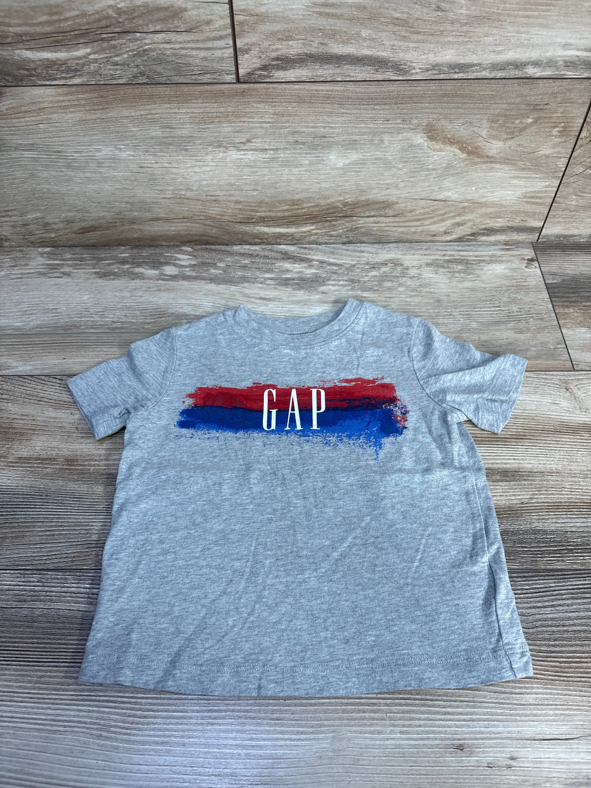 Front view of a grey Baby Gap short sleeve shirt showcasing a GAP logo over red and blue stripes on a wood-grain background