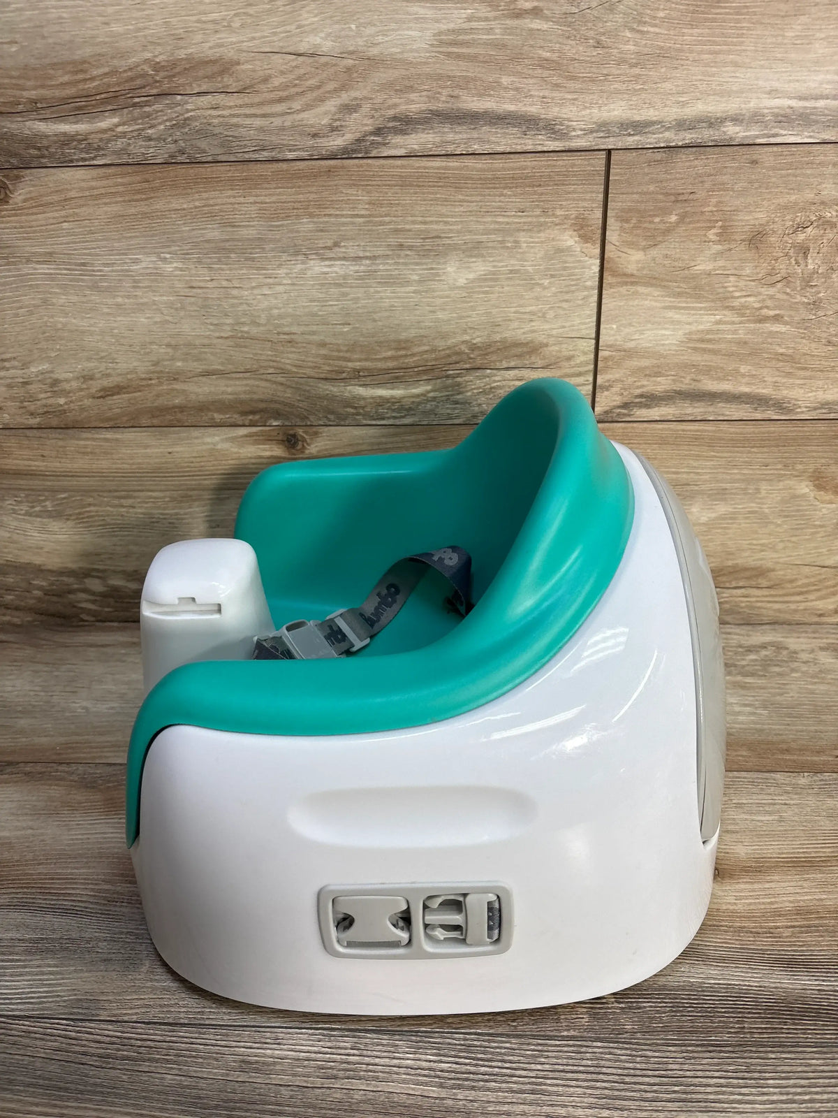 Front three-quarter view of Bumbo Multi Seat showcasing its teal seat, white base, and safety harness on a wood floor