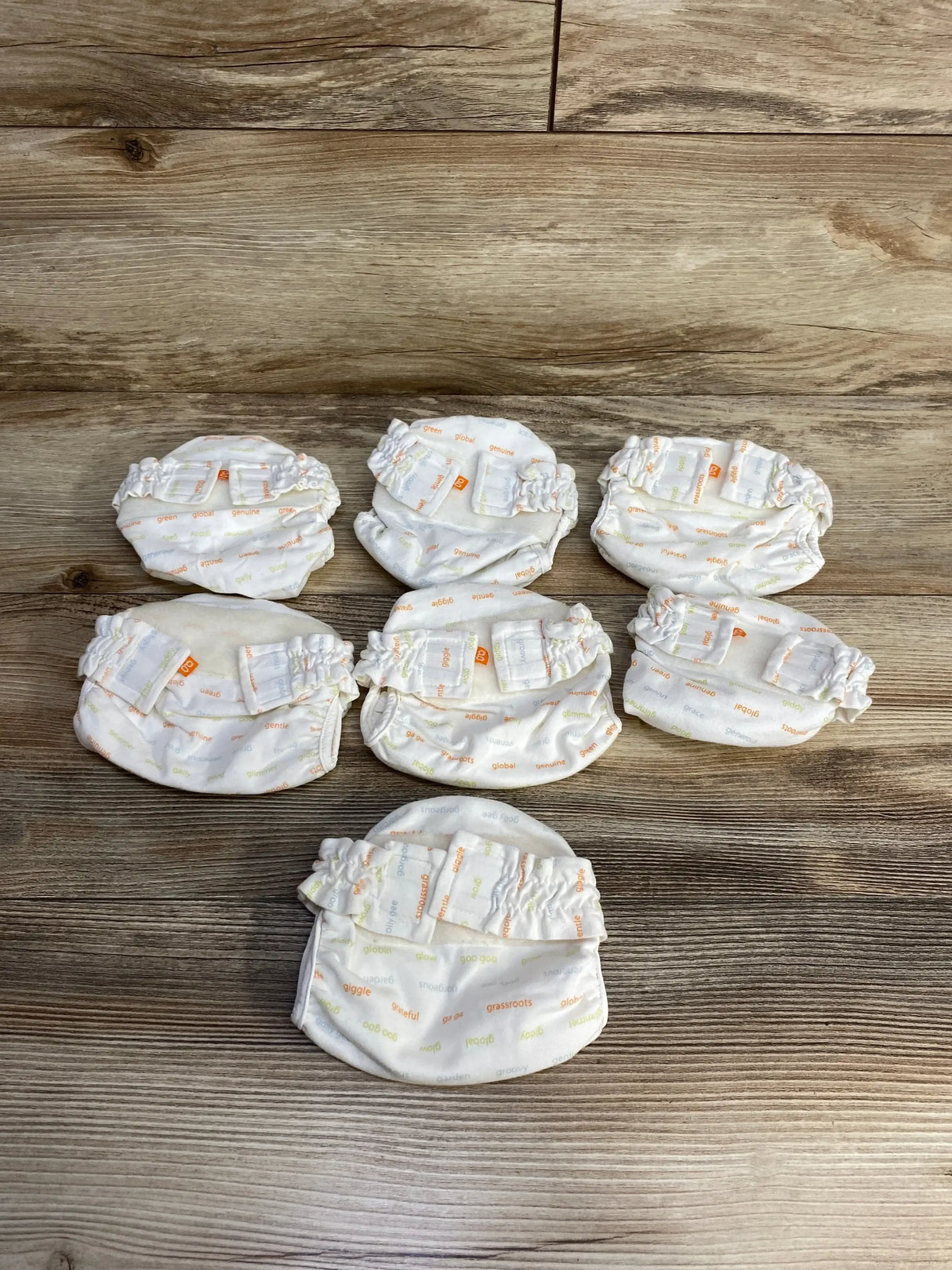 Top view of seven gDiapers newborn cloth diaper covers in ivory with multicolor word print and Velcro tabs on wood background