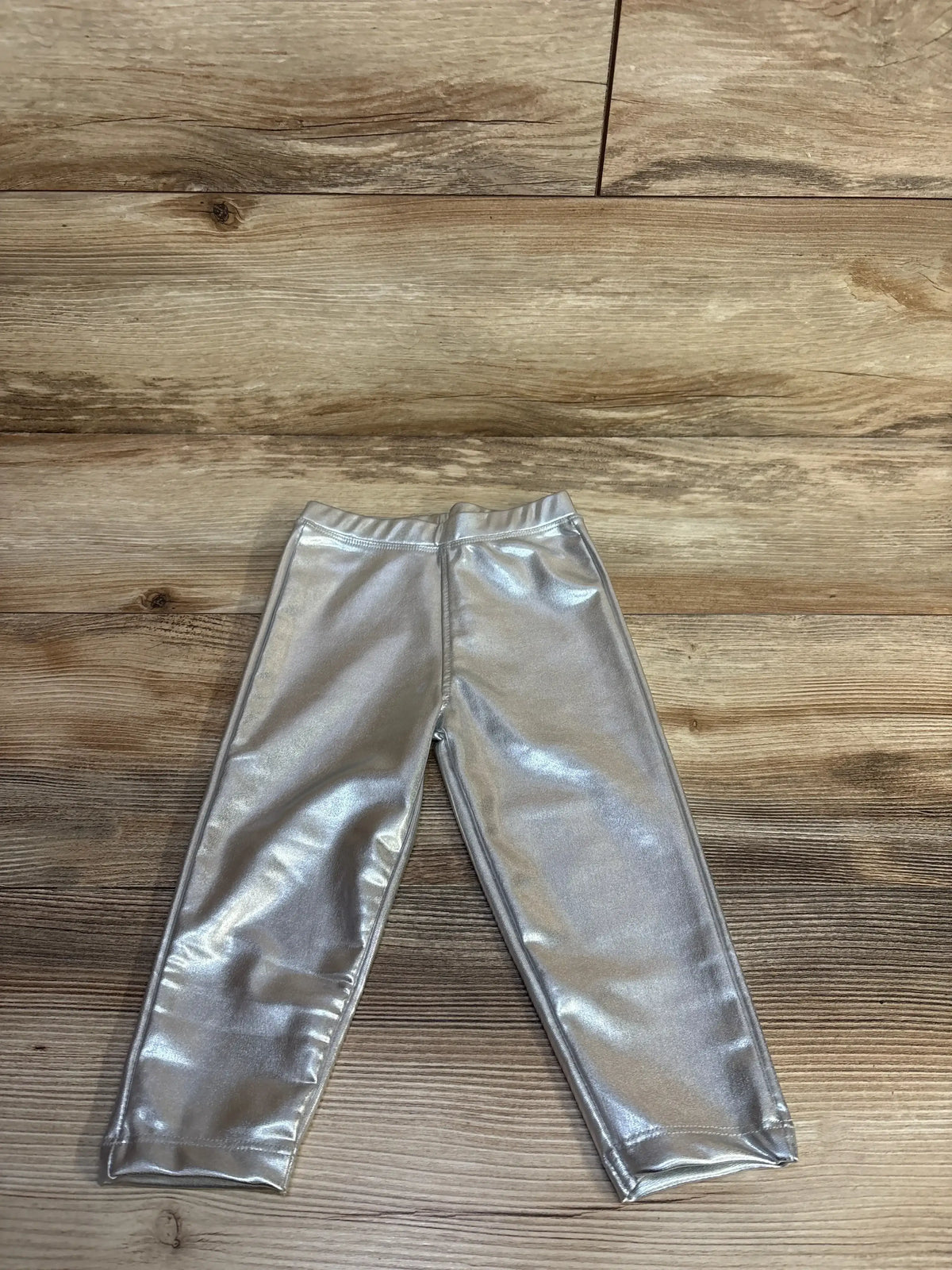 Flat lay of toddler silver metallic leggings showing shiny stretch fabric and elastic waistband on wood floor
