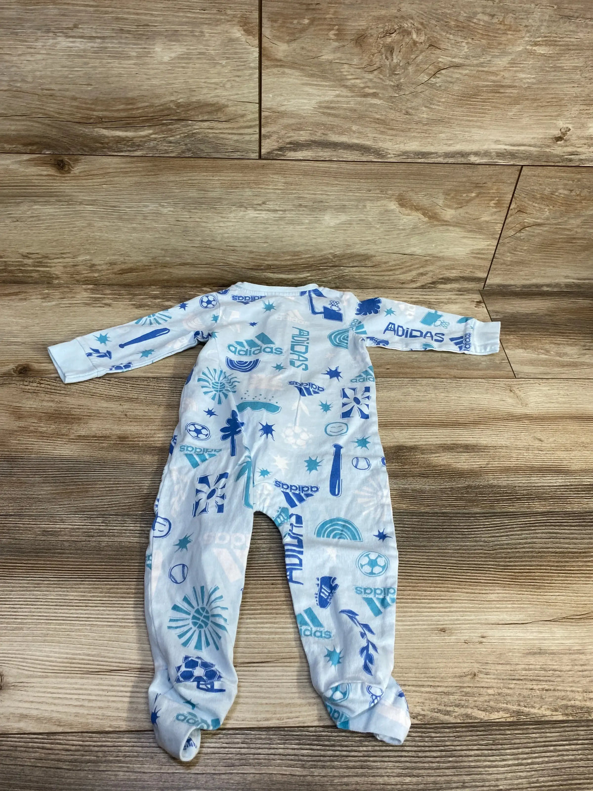 Flat lay of blue Adidas footed baby sleeper showcasing all-over Adidas logos and sports motifs, size 6 months, on wood floor