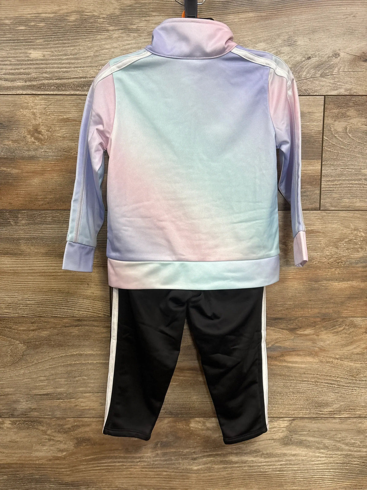Back view of Adidas toddler tracksuit, pastel purple jacket and black pants with white side stripes on wooden background