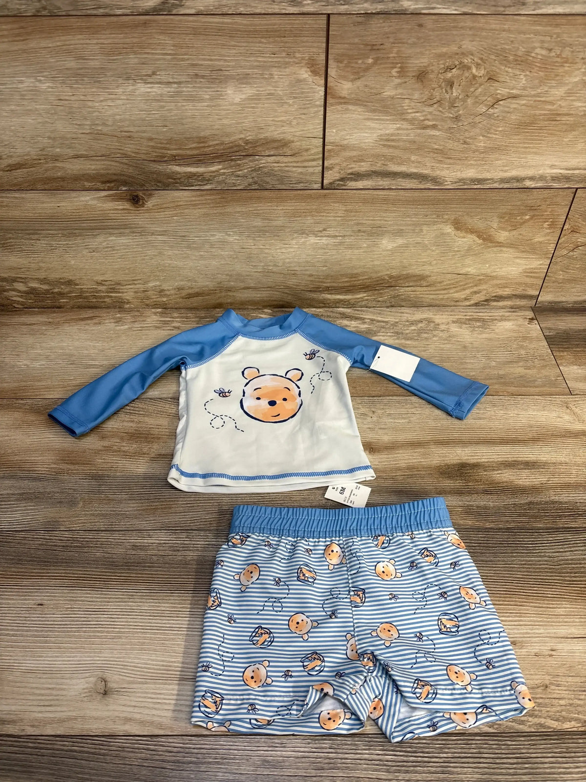 Flat lay of Disney Baby 2-piece Winnie the Pooh rash guard swim set, long-sleeve white and light-blue top with Pooh face and matching blue-striped shorts, size 6M, on wood floor