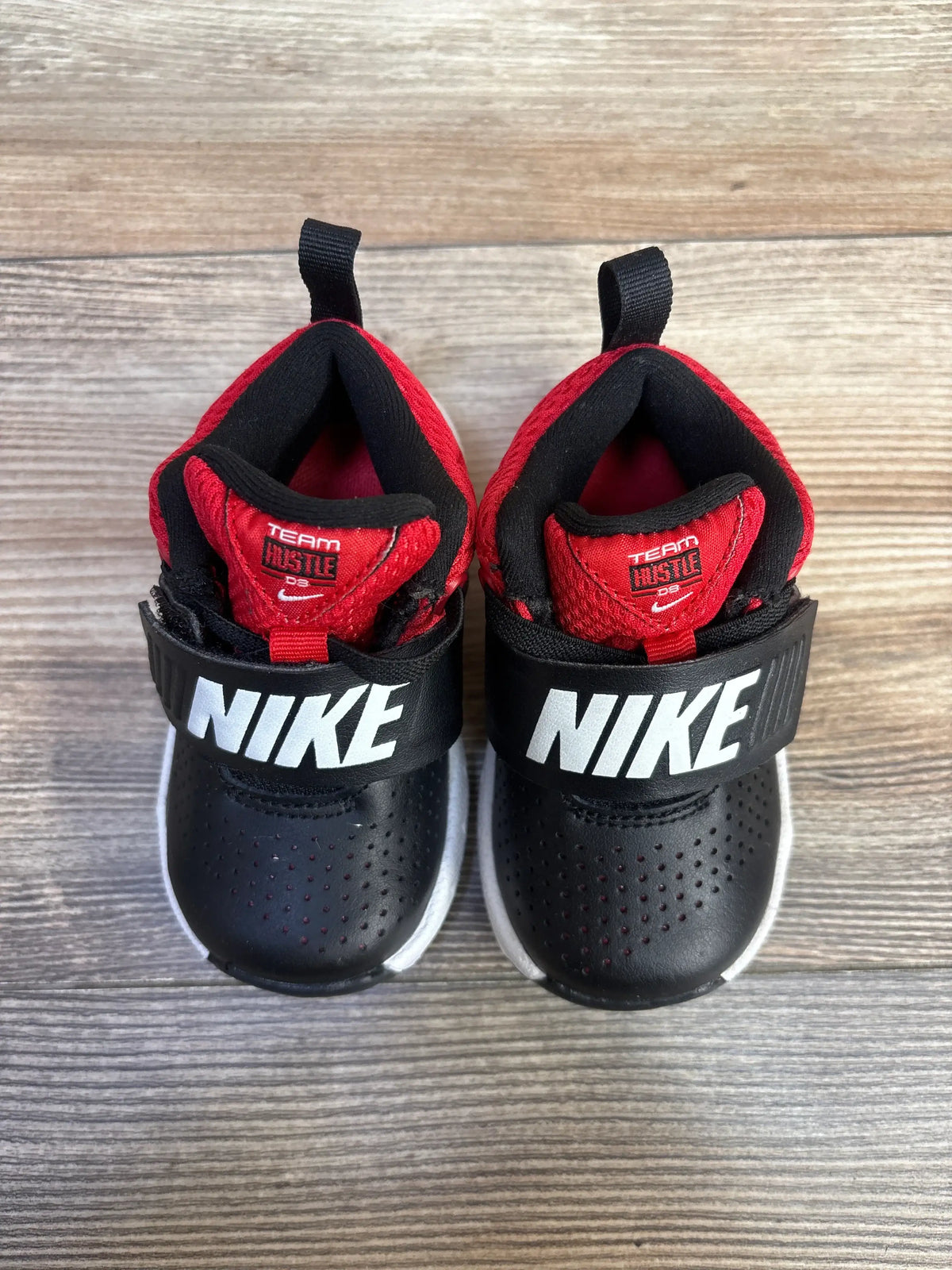 Front view of Nike Team Hustle D8 sneakers in black and university red with Nike straps and perforated toes on wood