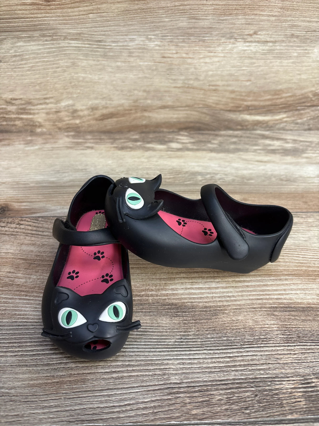 Black children's Mary Jane shoes with green-eyed cat face and pink paw print insoles on wooden surface