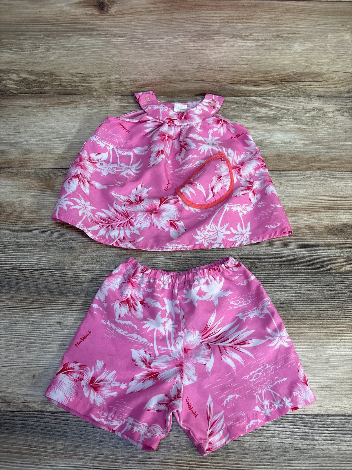 Pink floral Hawaiian toddler outfit set with matching top, shorts, and accessory on wood background