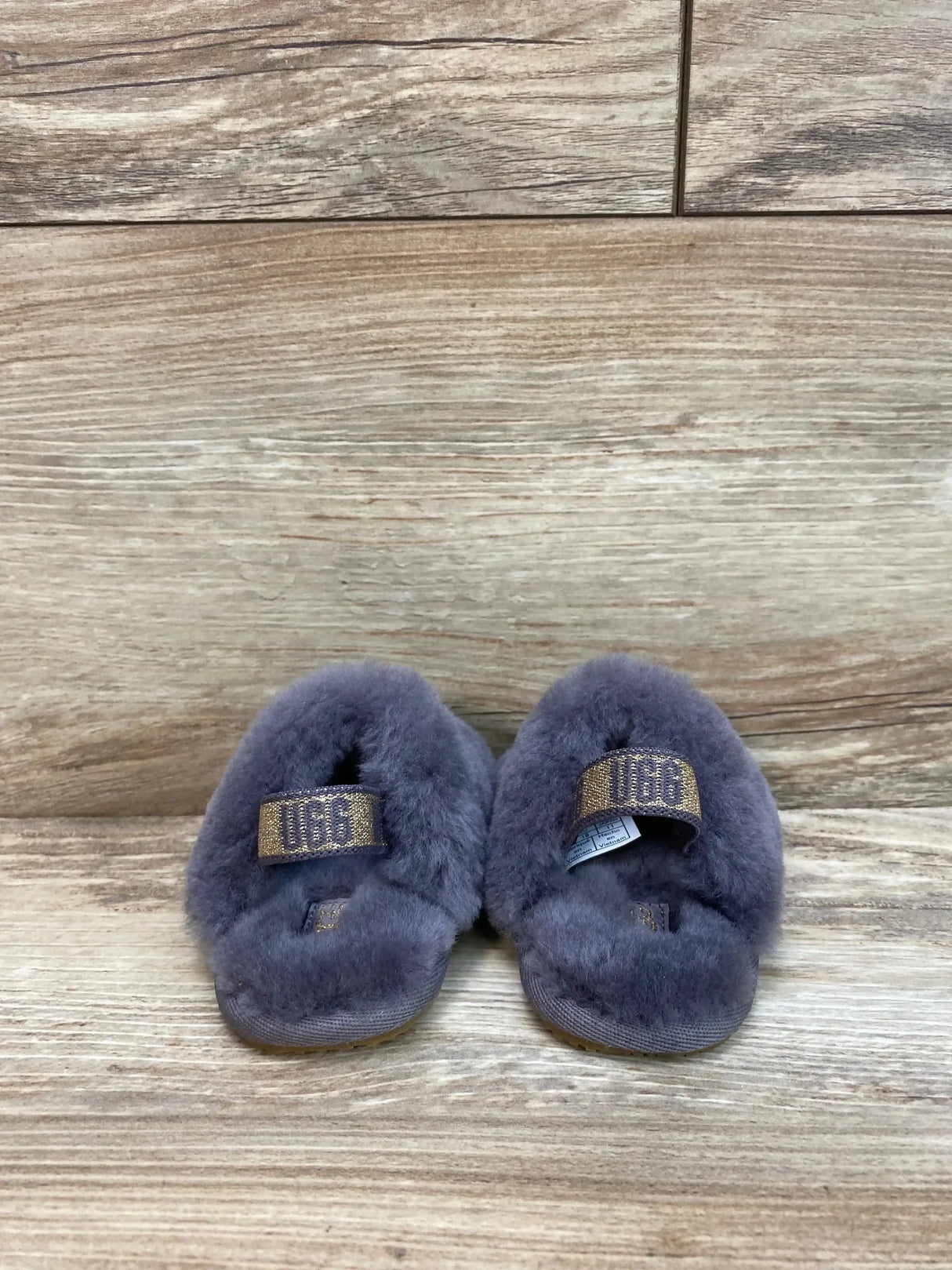 Back view of UGG Fluff Yeah Slides in grey showcasing fluffy sheepskin strap with UGG logo patch on wood plank floor