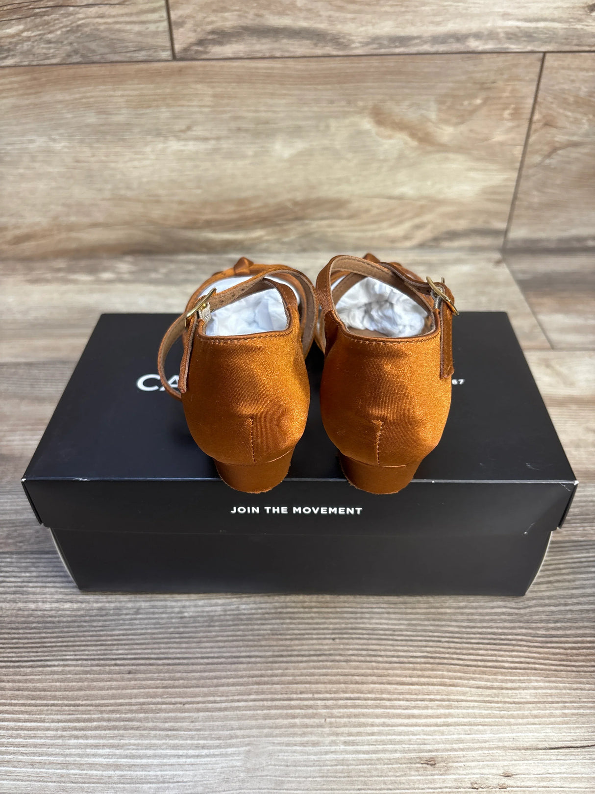 Rear view of Capezio Valentina ballroom flats in cinnamon satin showing ankle straps and heels on black shoebox, wood floor