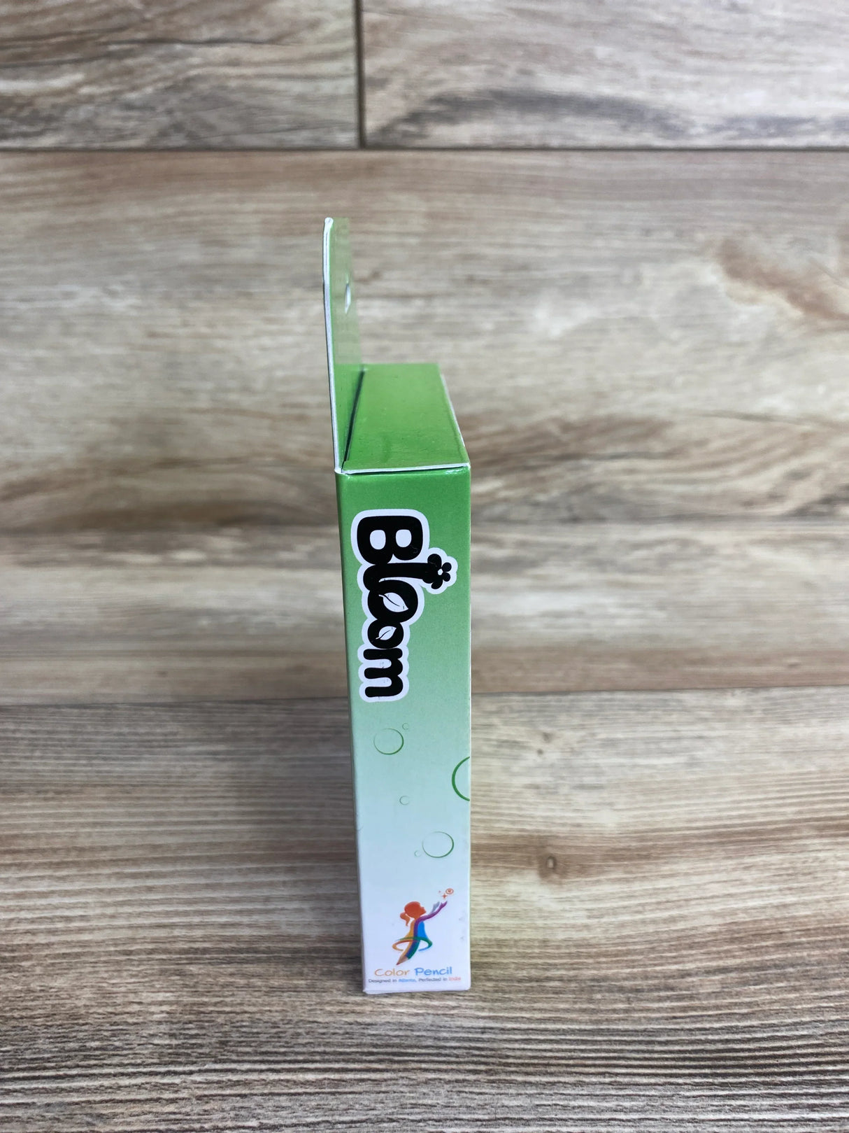 Side view of Bloom Color Pencil box showcasing green gradient packaging with brand logo on wood plank floor