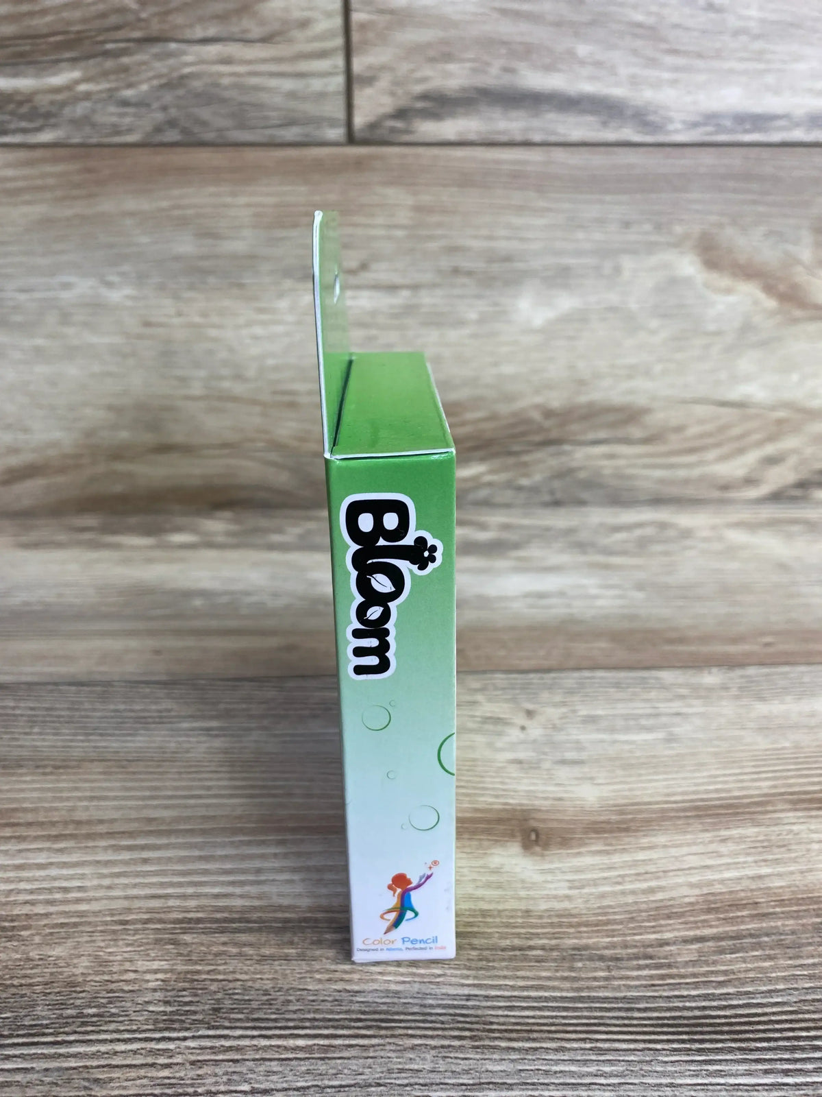 Side view of Bloom Color Pencil box showcasing green gradient packaging with brand logo on wood plank floor