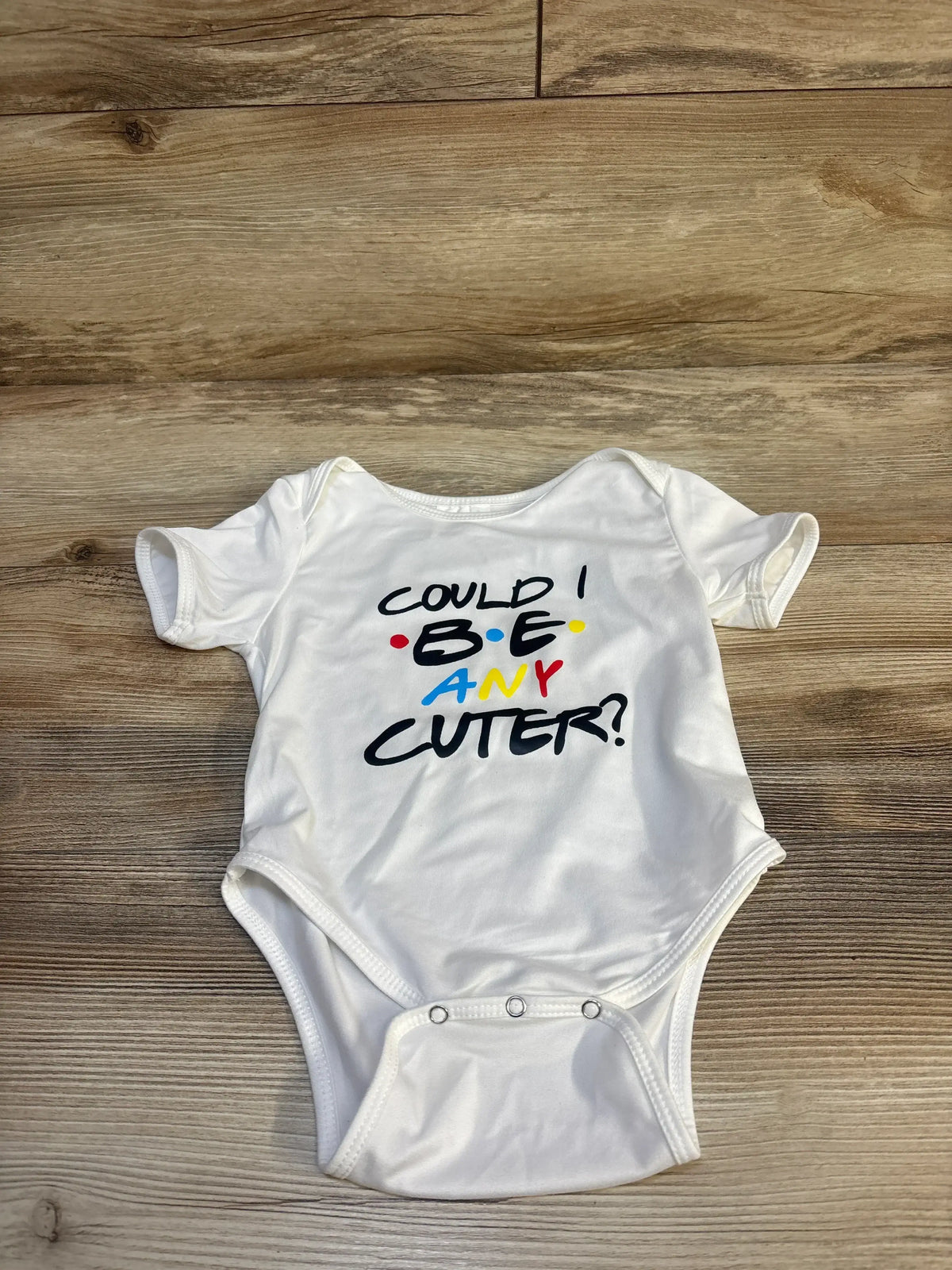 White baby onesie with colorful text 'Could I BE any cuter?' on wood floor background