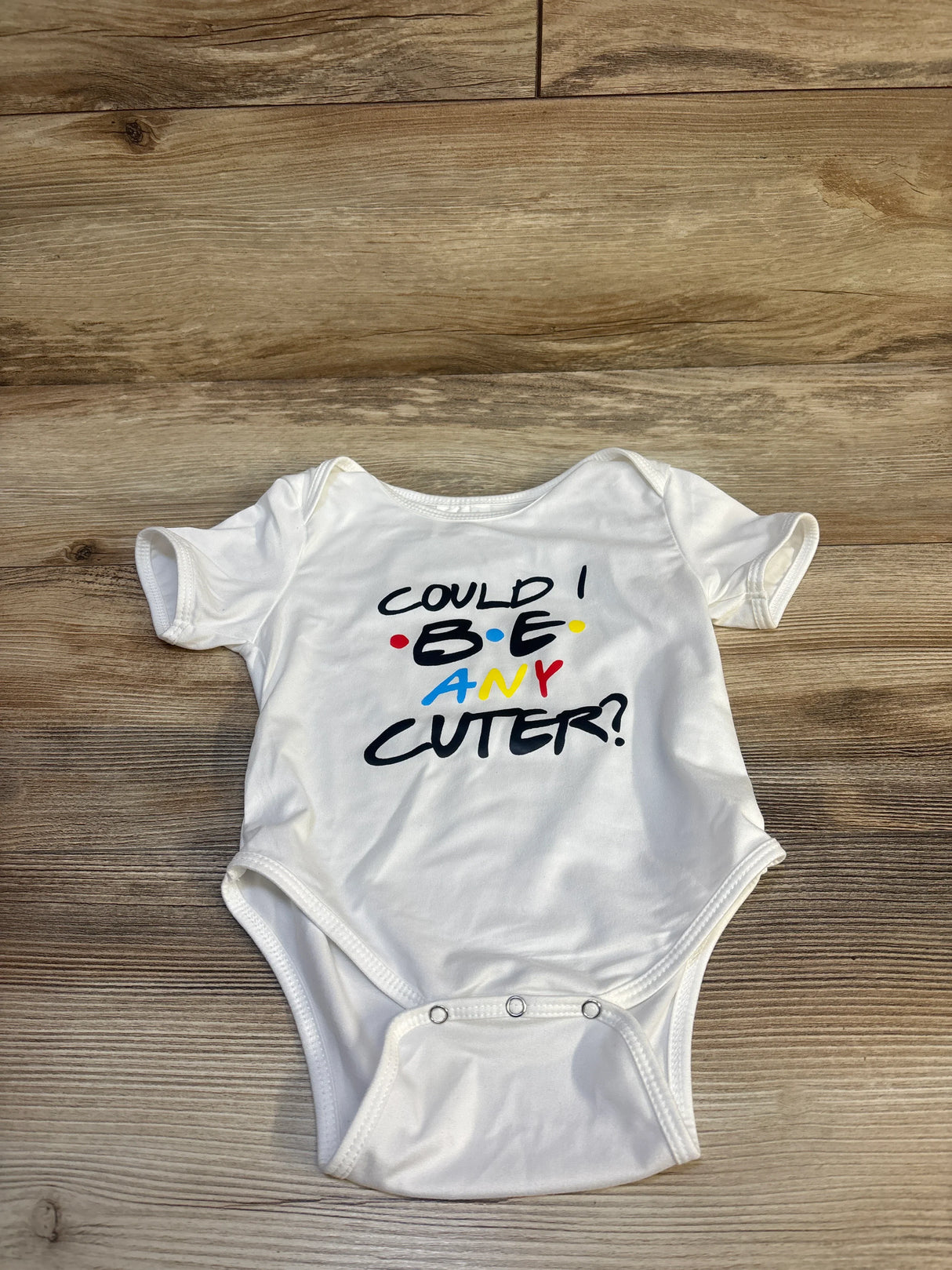 White baby onesie with colorful text 'Could I BE any cuter?' on wood floor background