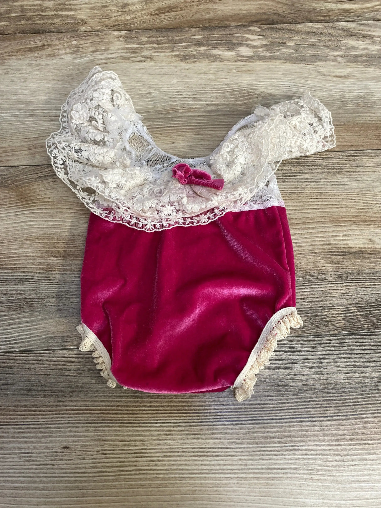 Flat lay newborn lace velvet romper in deep pink with white lace yoke, bow, and ruffled leg trim on wood background