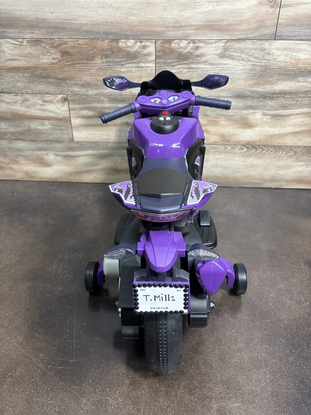 Front view of purple Marvel Black Panther ride-on motorcycle showing training wheels and a custom T. Millz Wakanda license plate with wood-paneled background