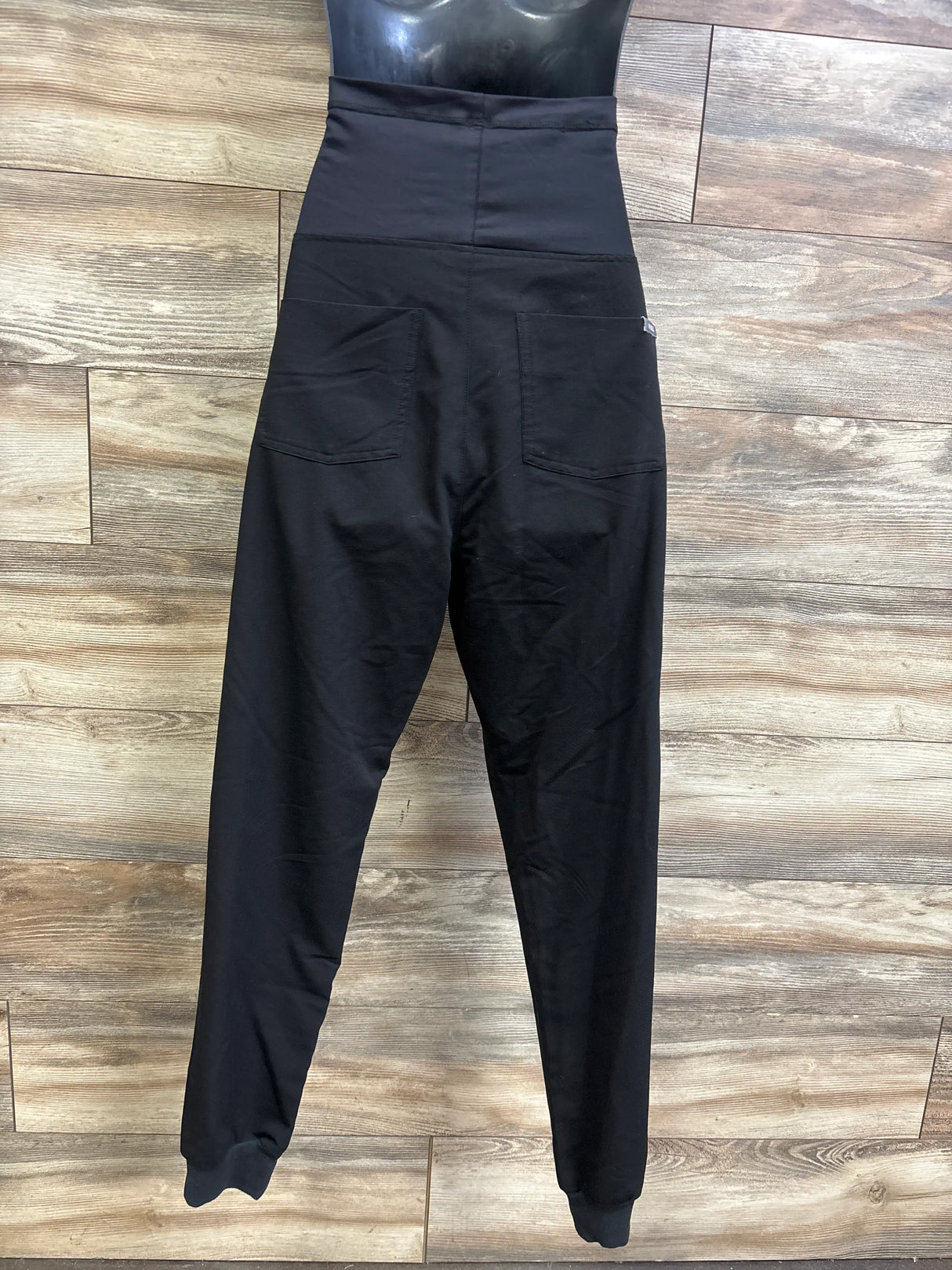 Back view of FIGS maternity scrub pants, black, high over-belly panel, two back pockets, cuffed ankles, wood backdrop