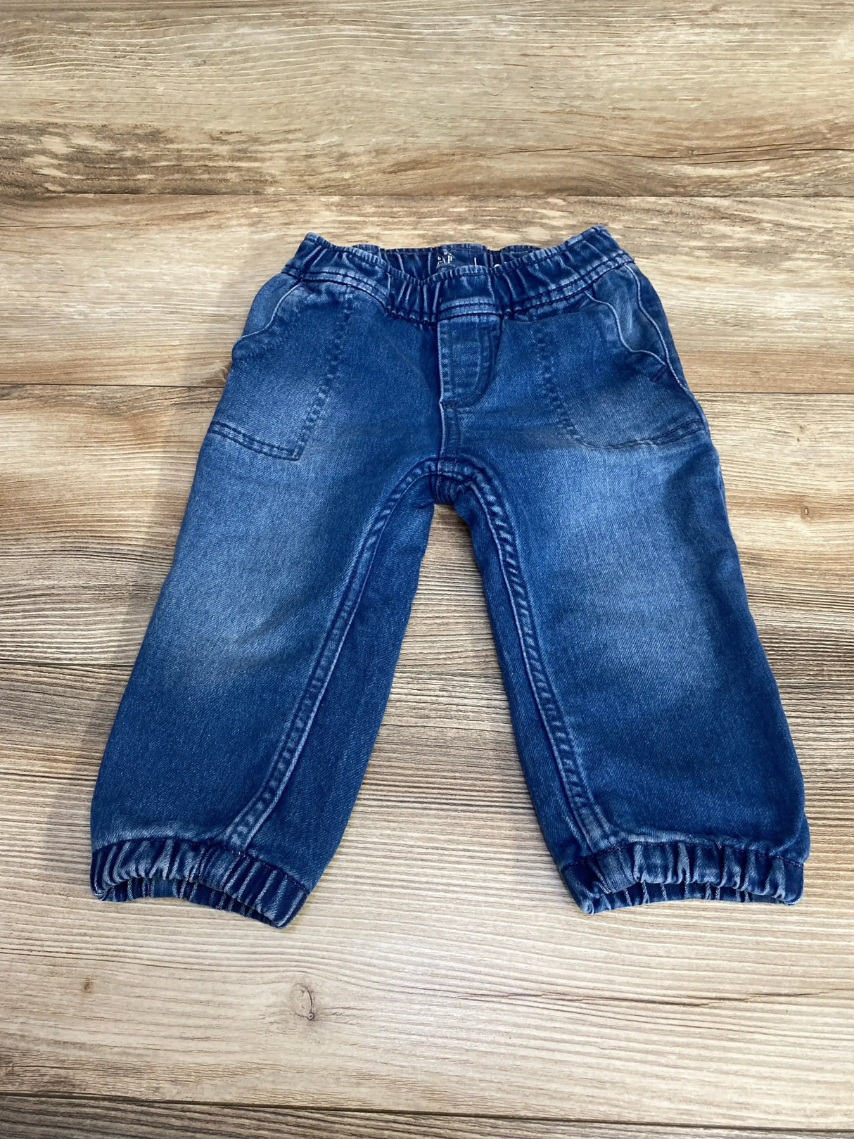 Flat lay of Baby Gap blue denim jogger jeans showcasing elastic waistband and cuffed ankles with whiskered wash on wood plank floor