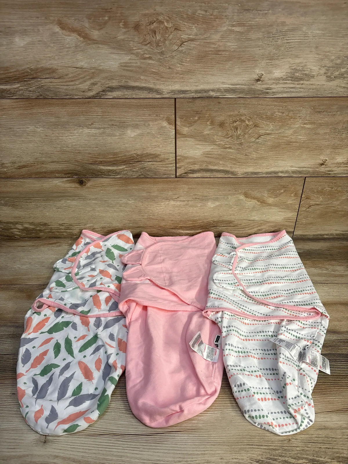 Top-down view of three Gilquen swaddle wraps (3–6 months) showing feather print, solid pink, and dotted stripe designs on a wood background