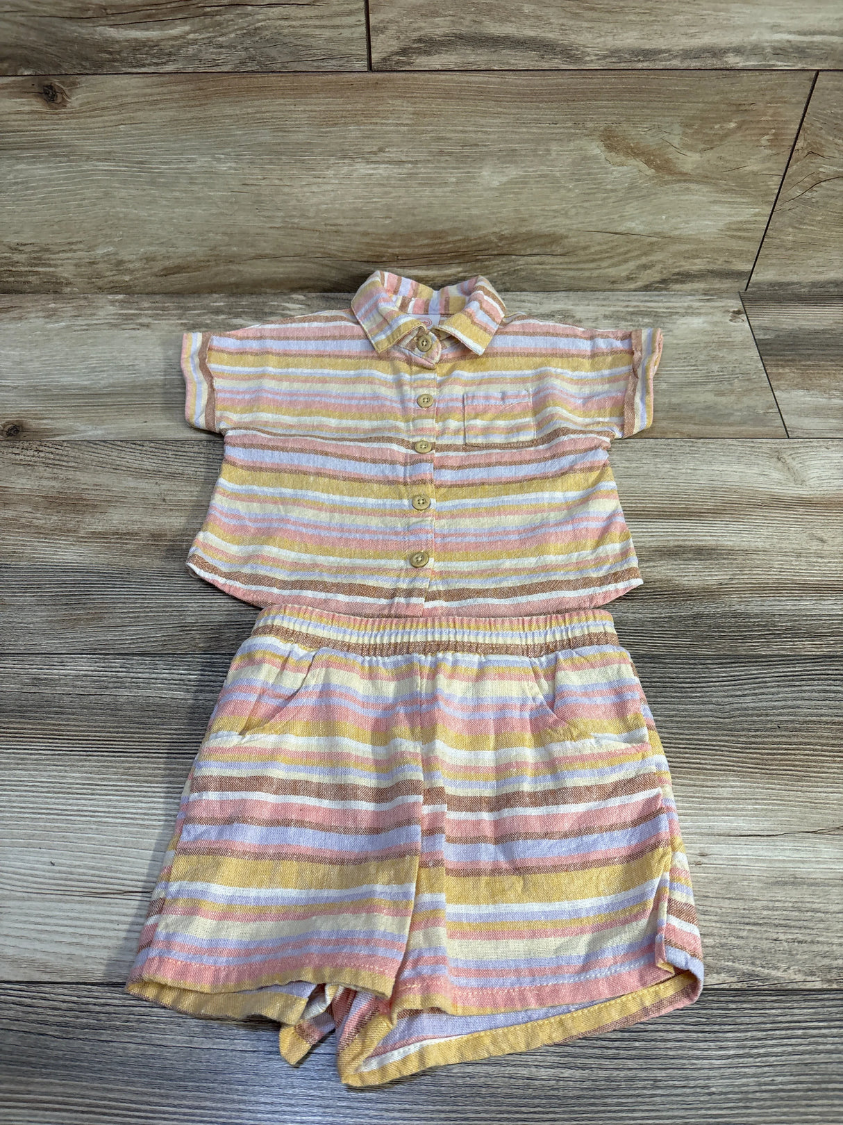 Flat lay of toddler yellow-pink striped linen-blend button-up shirt and shorts showing front buttons and pockets on wood floor